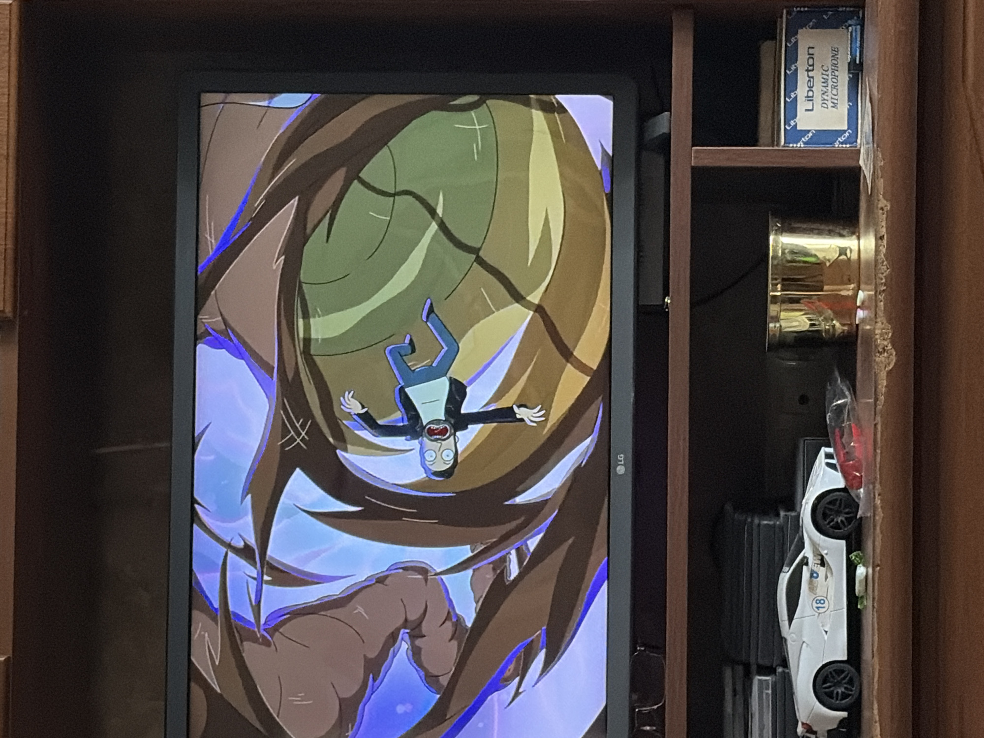 In an indoor setting, a television displays a scene from an animated show, possibly "Rick and Morty." A character resembling Rick Sanchez is depicted falling or being pulled through a swirling, abstract vortex of brown, green, and blue colors. Below the television, on a wooden shelf, sits a white toy sports car with the number "18" on its side. To the right of the car, there is a golden cup and a blue box labeled "Liberton DYNAMIC MICROPHONE." The overall scene appears to be in a domestic environment. The location is Unknown, Unknown.