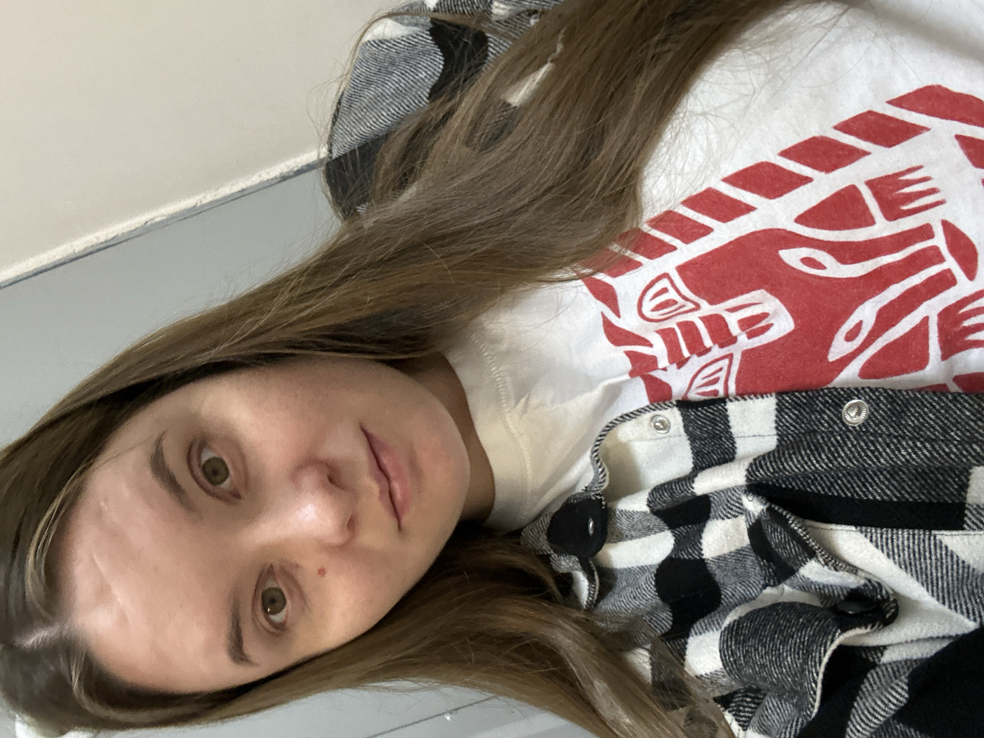 A young woman with long brown hair is taking a selfie. She is wearing a white t-shirt with a red graphic design and a black and white plaid shirt over it. She is indoors, in what appears to be a hallway, with a grey wall behind her. There are no visible indications of her location in Chaikovsky, Russia.