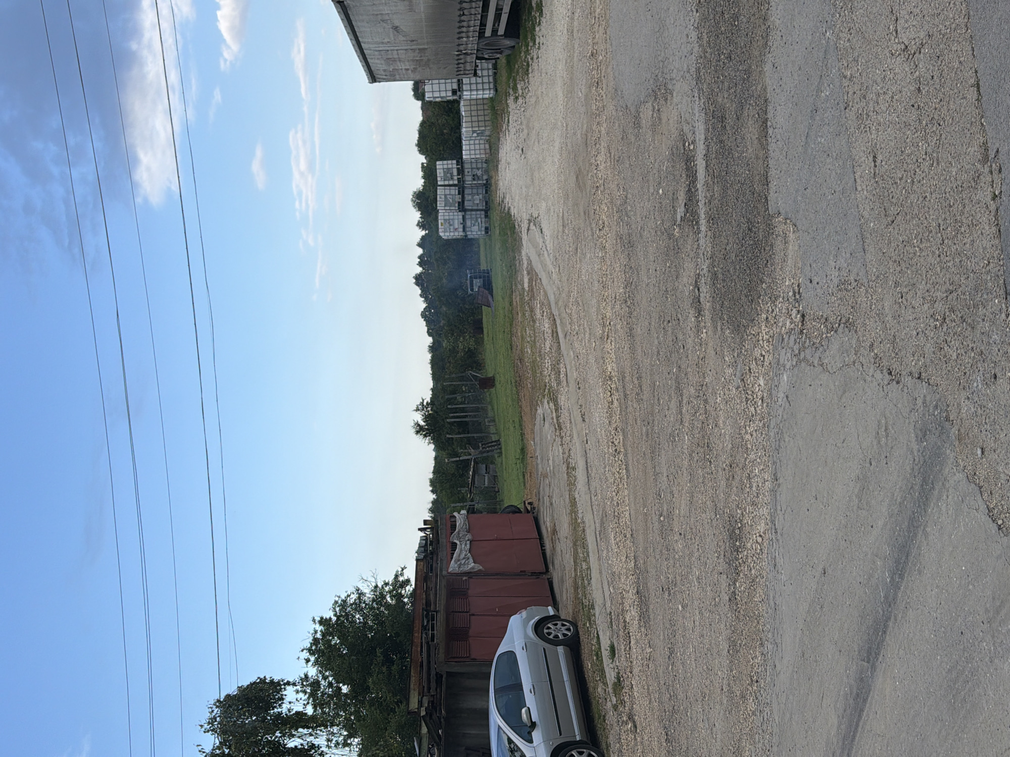 The image depicts an outdoor scene in Chervena Voda, Bulgaria. In the foreground, a cracked asphalt road transitions into a gravel-covered lot. To the left, a silver sedan is parked beside a red garage with two large, double doors. The garage is constructed from metal sheeting and appears weathered, with a tarp draped over its roof. Behind the garage, several rusted metal structures are stacked.
Further into the scene, the gravel lot gives way to a grassy area. In the middle ground, a collection of large, white plastic containers, possibly IBC totes, are stacked in multiple rows. To the right of these containers, the rear of a large, grey truck trailer is visible.
The background consists of a treeline and a bright blue sky with scattered clouds. Several power lines are strung across the upper portion of the image. The overall impression is of an industrial or storage yard in a rural or semi-rural setting. No people are visible in the image.