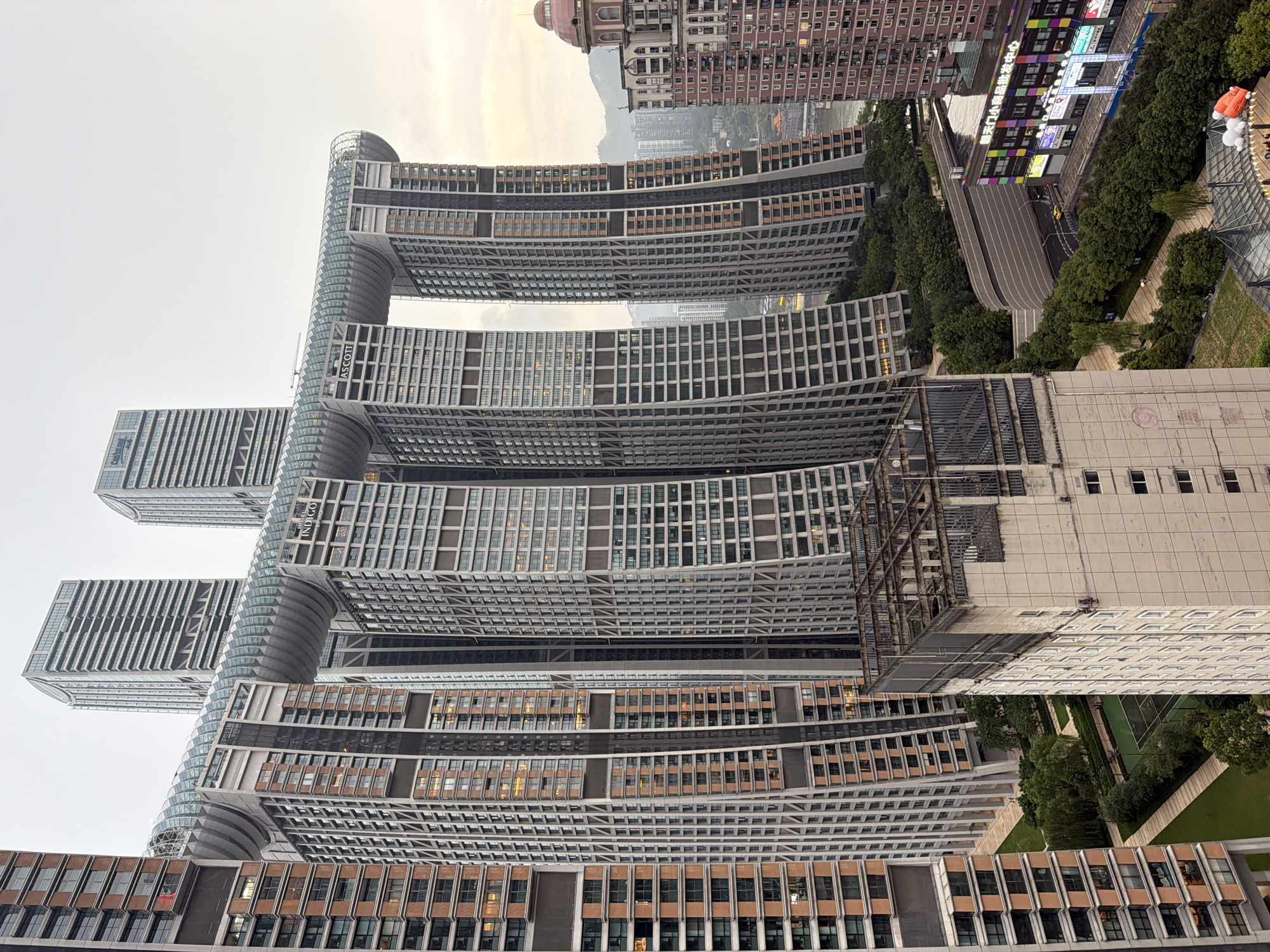 The image depicts the Raffles City Chongqing complex in Yuzhong District, China, a striking architectural marvel. The complex features six interconnected skyscrapers topped by a horizontal, crystal-like structure named "The Crystal." The buildings are densely packed, showcasing a modern urban landscape with a mix of residential and commercial spaces. The sky is overcast, hinting at either dawn or dusk. In the foreground, a concrete structure with exposed metal beams suggests ongoing construction or a disused rooftop. Below, manicured green spaces and a tennis court provide a contrast to the imposing skyscrapers. Further in the background, other buildings and distant mountains are visible, suggesting the complex is situated within a larger metropolitan area. The architecture is characterized by its geometric shapes, cantilevered sections, and the prominent use of glass and metal. The overall impression is one of grand scale and contemporary urban design.