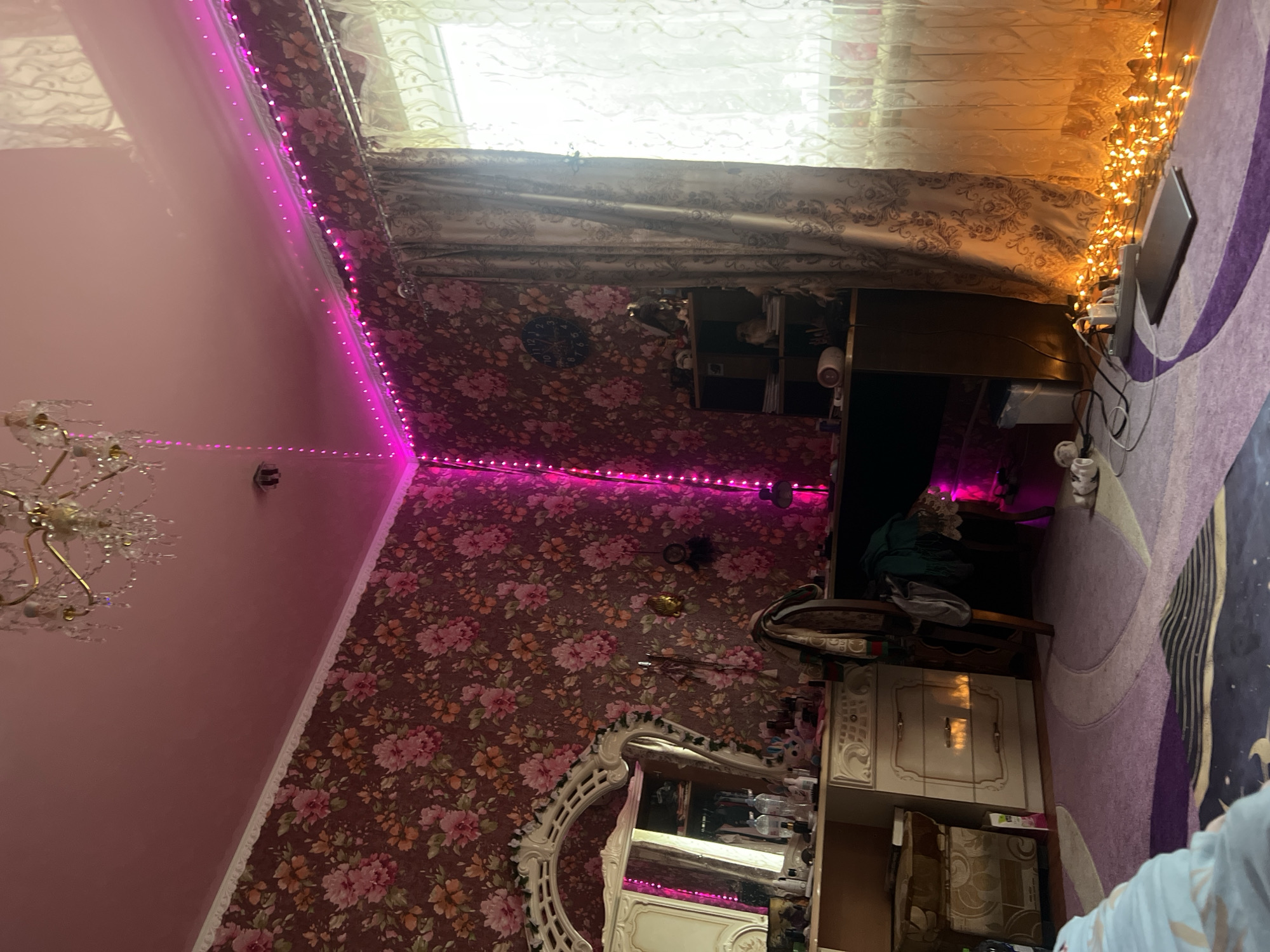 The image shows a dimly lit bedroom with floral wallpaper and pink LED strip lights illuminating the ceiling. A crystal chandelier hangs from the ceiling. A large ornate mirror with a carved frame is positioned against the floral wallpaper. Below the mirror is a white dresser with several drawers. To the right of the dresser is a dark shelf unit with various items on it, including a clock. A window covered by patterned curtains is to the far right, with a radiator beneath it. The floor is covered by a patterned rug in shades of purple and blue. A laptop, an extension cord with plugs, and a string of orange fairy lights are on the floor near the window. Clothing is draped over a chair. The overall impression is that of a personalized and somewhat cluttered bedroom space. There are no people visible in the image.