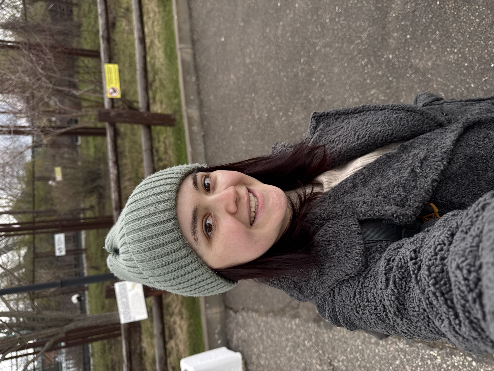 A young woman with dark hair and brown eyes takes a selfie outdoors in Izhevsk, Russia. She smiles broadly, showing her teeth, and appears happy. She is wearing a light green knitted beanie and a thick, dark grey, textured coat, suggesting cool or cold weather.
The setting is an outdoor area, likely a park or similar recreational space. In the background, there is a wooden fence or railing. Beyond the fence, grass and bare tree branches are visible, indicating an early spring or late autumn season. The ground in the foreground is paved, possibly asphalt, with a slightly textured appearance.
Attached to the wooden fence are a few signs. One prominent yellow rectangular sign features a prohibition symbol (a black circle with a red diagonal line) over what appears to be a dog icon, likely indicating "no dogs allowed" or a similar restriction. There is also a white rectangular sign with illegible text. A white, possibly plastic, object rests on the ground in the lower portion of the image. The lighting is soft and even, suggesting an overcast day.