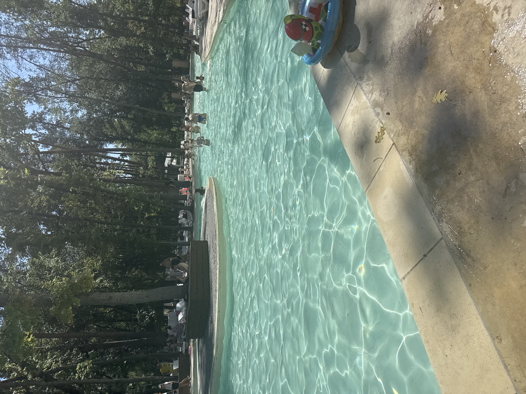 A poolside scene at a water park in Orlivshchyna, Ukraine, is depicted. In the foreground, the light blue, rippling water of a swimming pool dominates the lower half of the frame. The water is illuminated by sunlight, creating shimmering reflections on its surface. To the right, a tan-colored concrete edge of the pool is visible, with a small inflatable toy boat carrying a watermelon and a duck floating near the edge. In the background, a crowd of people are enjoying the pool, with some swimming and others wading. Many individuals are wearing swimwear. Beyond the pool, a lush green forest with tall trees forms a dense backdrop, suggesting the water park is situated within a natural, wooded environment. The sky is a clear blue, indicating a sunny day.