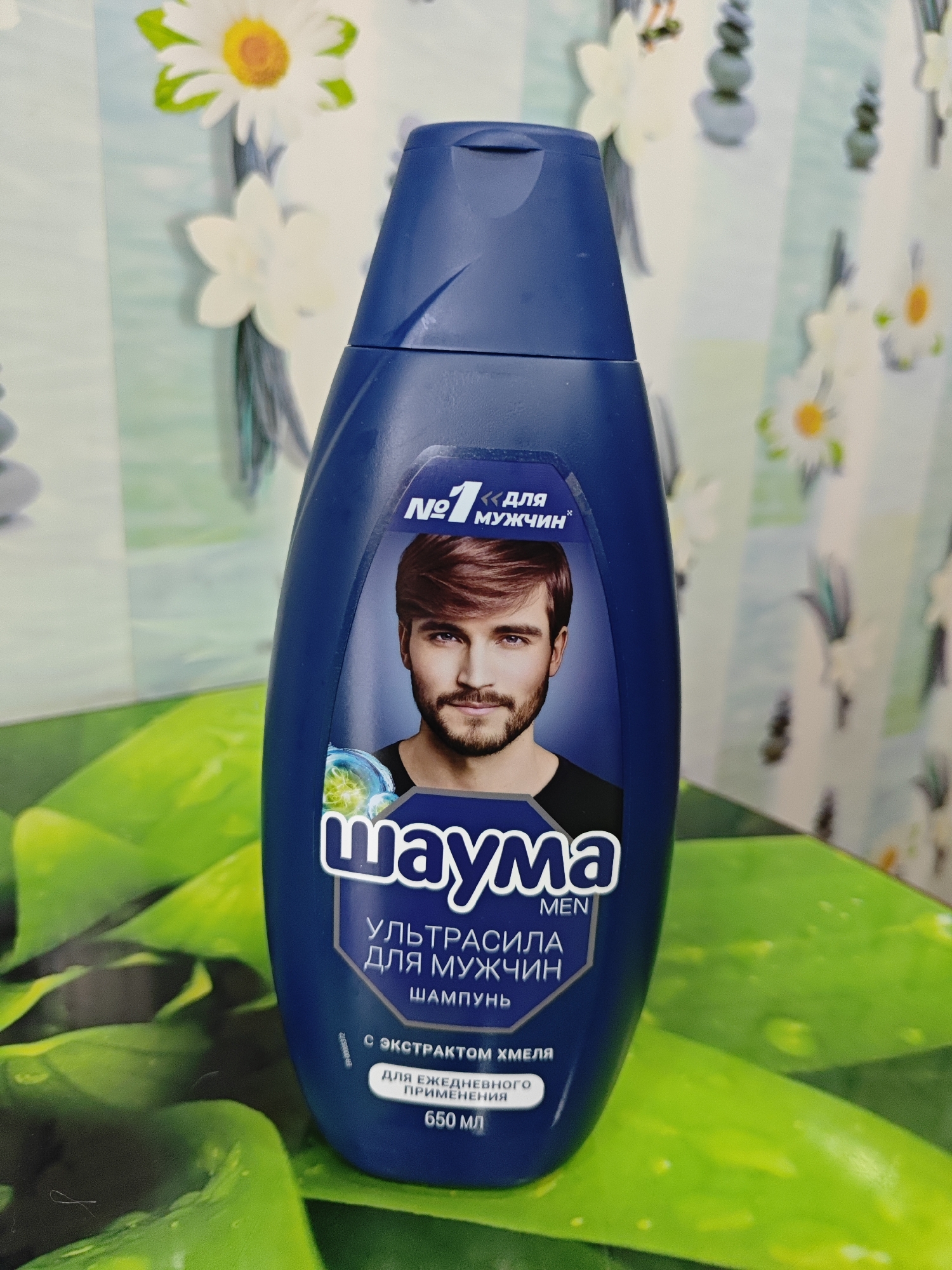 A dark blue plastic bottle of shampoo is positioned in the foreground, angled slightly to the left. The bottle features a prominent label with white text and graphics. At the top of the label, a stylized "No. 1" is followed by Russian text that reads "для мужчин" (for men). Below this, a portrait of a man with short, reddish-brown hair and a beard is displayed. The brand name "шаума" (Shauma) is written in large white letters with a blue outline. Beneath the brand name, Russian text indicates "УЛЬТРАСИЛА ДЛЯ МУЖЧИН" (Ultrasila for Men) followed by "ШАМПУНЬ" (Shampoo). Further down, there is text stating "С ЭКСТРАКТОМ ХМЕЛЯ" (with hop extract) and "ДЛЯ ЕЖЕДНЕВНОГО ПРИМЕНЕНИЯ" (for daily use). The volume "650 мл" (650 ml) is also present. The bottle is placed on a surface with a bright green, leafy pattern, possibly a placemat or tablecloth. The background is out of focus, showing a light blue and white striped pattern with floral motifs, resembling wallpaper or a curtain. The overall lighting is bright, highlighting the shampoo bottle. The image does not provide any information or context related to the city of Polyarny, Russia.