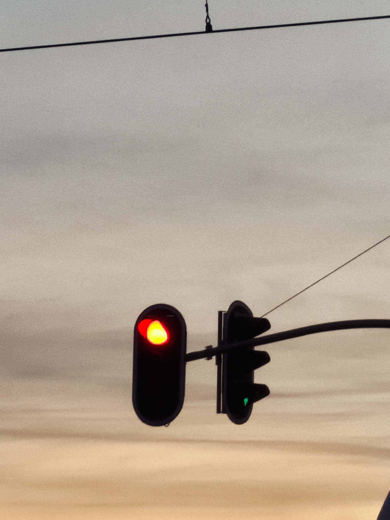 The image shows a traffic light signal with a red light illuminated in the top signal, and a green light illuminated in the bottom signal. The traffic lights are suspended over a street, with power lines visible in the background. The sky is overcast and appears to be at dusk. The scene is likely located in the city of Gdańsk, Poland, as indicated by the presence of traffic lights and power lines commonly found in urban environments. There are no people or vehicles visible in the image.