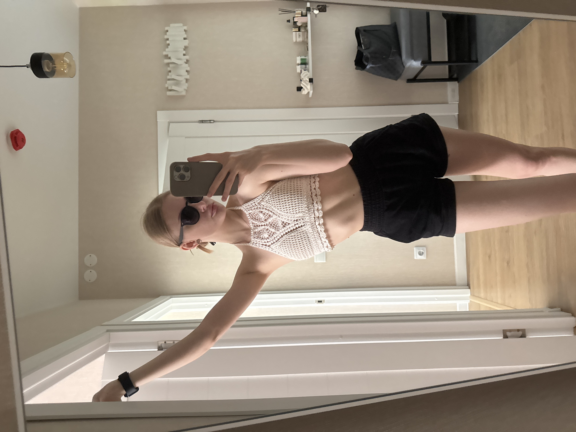 A young woman is taking a selfie in a mirror. She is wearing a white crochet halter top and black shorts. She is also wearing sunglasses and a black watch. The background includes a room with a light fixture, a shelf with various items, a chair, a door, and an ornate white wall decoration. The floor is light-colored wood. Based on visual cues such as architectural details and light quality, it is highly unlikely that the location is in the city of Perm, Russia. The environment lacks characteristics typical of that region.