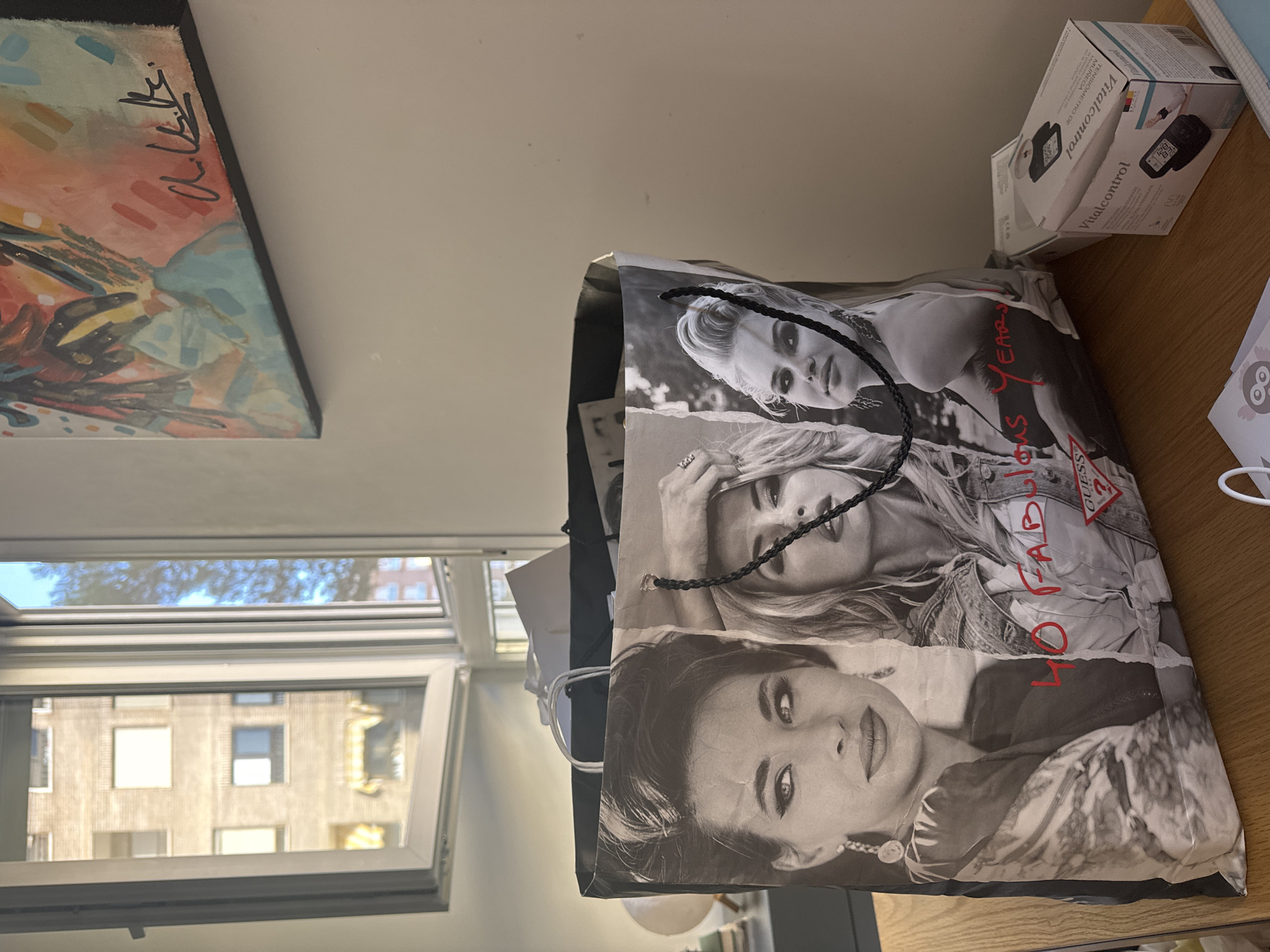 This image depicts an indoor scene, likely a room in a building. In the foreground, a large Guess shopping bag is prominently featured, adorned with black and white images of women and the text "40 FABULOUS YEARS". The bag is resting on a wooden surface, possibly a desk or table. To the right of the bag, a small white box labeled "Vitalcontrol" is visible. In the background, a window offers a view of an urban environment with multi-story buildings and trees. The lighting suggests it is daytime. The scene does not provide any specific visual cues to determine its location in the city of Murcia, Spain.