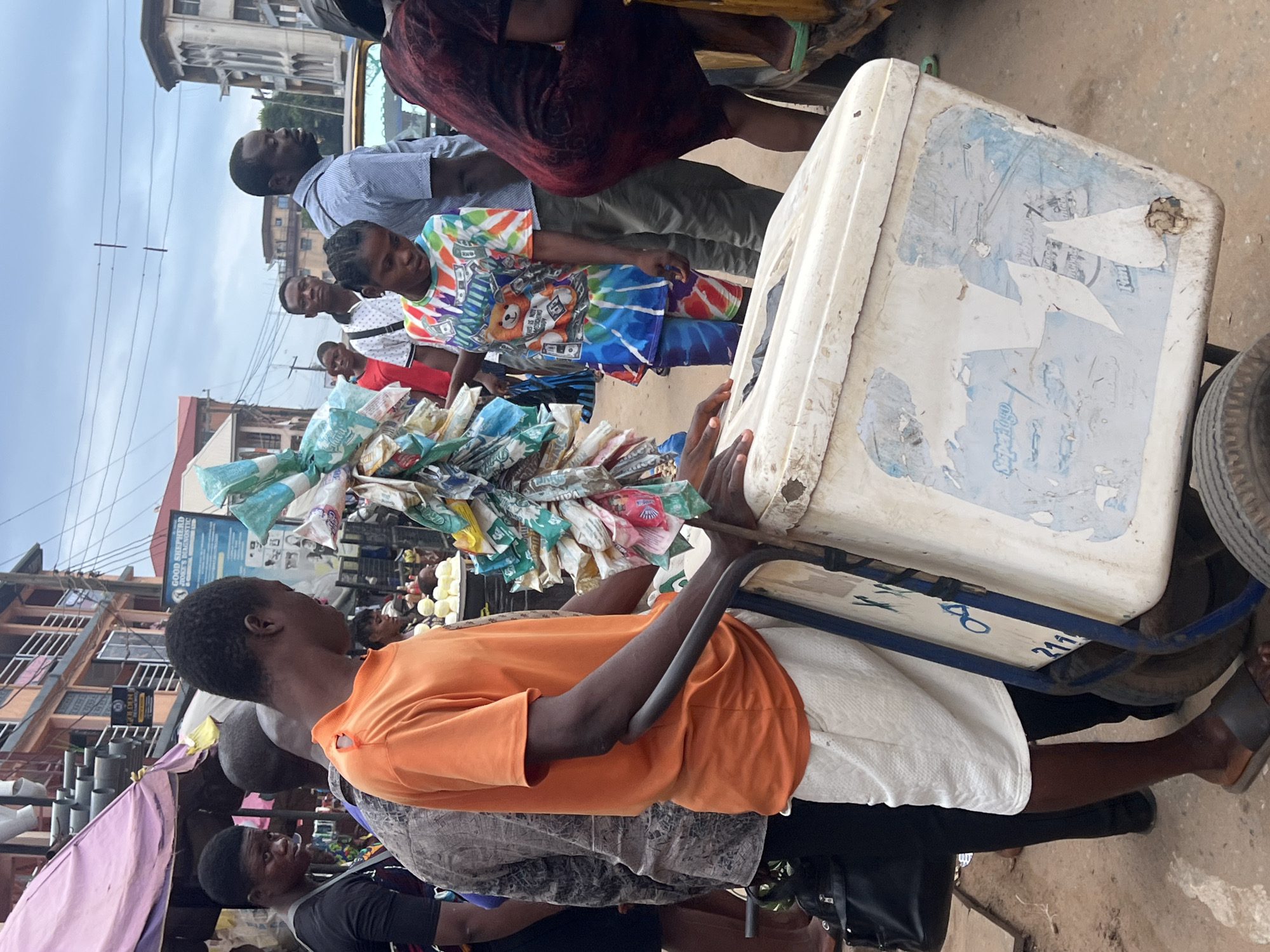 In Nkpor, Nigeria, a street scene unfolds with individuals engaged in commerce. A man in an orange t-shirt and white shorts pushes a white cooler on wheels, its surface worn with peeling stickers and blue markings. Beside him, a young woman wears a colorful tie-dye t-shirt adorned with cartoon imagery and money graphics, paired with matching patterned shorts. Several other people are visible in the background, including a woman in a dark patterned dress and a man in a blue striped shirt. The environment suggests a busy market or roadside setting, with buildings lining the street and various goods displayed under awnings. The lighting indicates it is daytime.