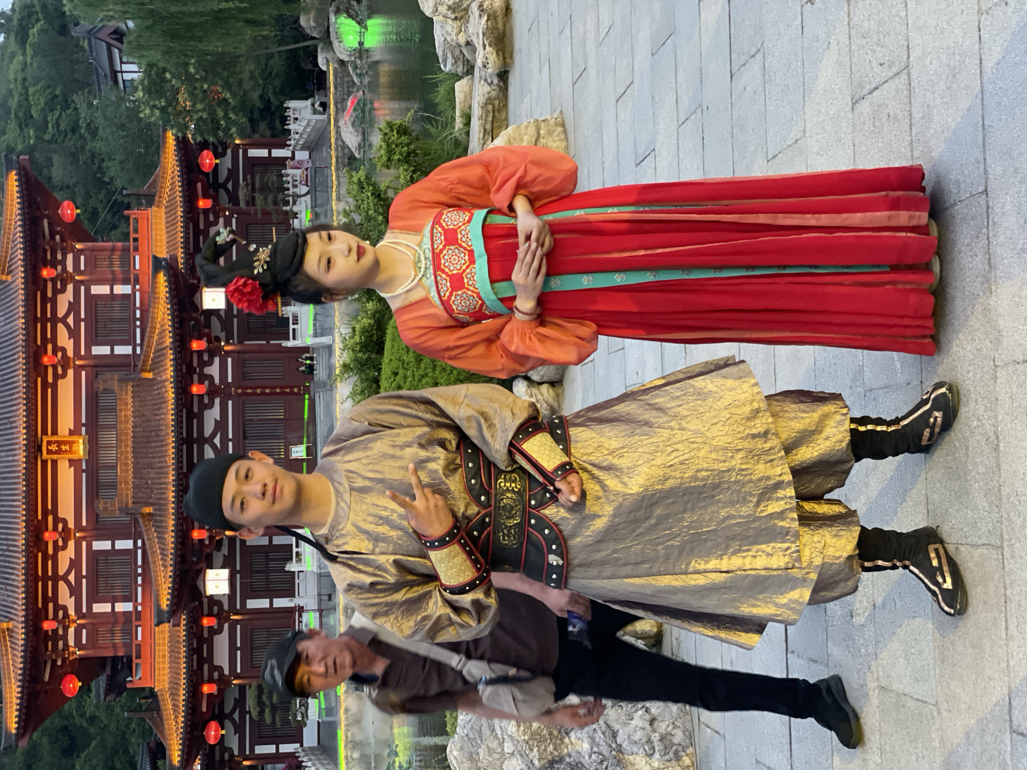 In Lintong District, China, a man and woman are dressed in traditional clothing and stand for a photograph near a pavilion with red lanterns. The woman wears a red and orange dress, and the man wears a gold garment. Another man stands nearby. A body of water and trees are visible in the background.