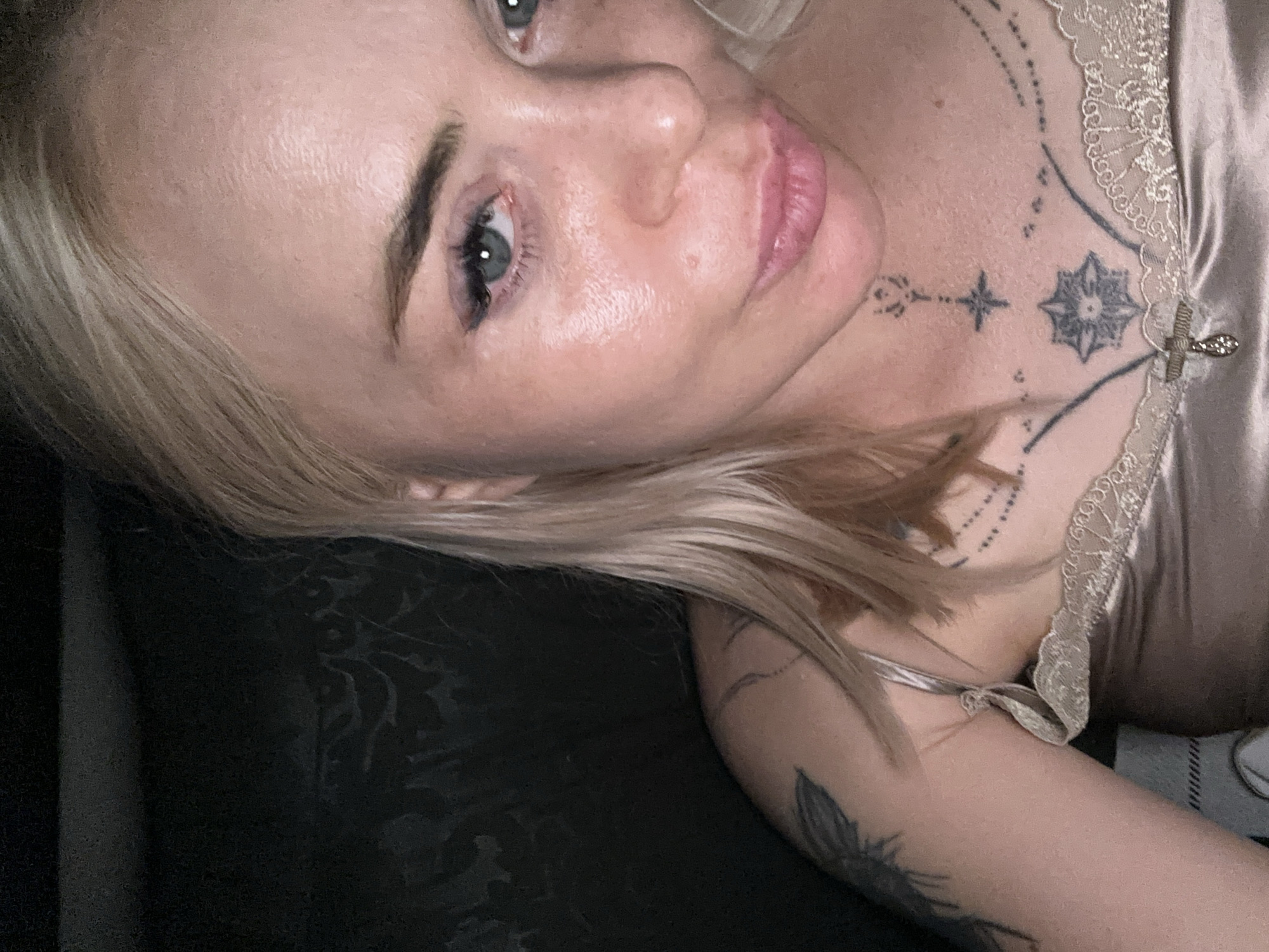 A close-up portrait depicts an adult female with blonde hair and light blue-green eyes, captured under direct frontal lighting that highlights the texture and sheen of her skin. She has a subtle smile. Visible tattoos include a dark, stylized design on her left upper arm/shoulder and a collection of geometric and linear patterns on her neck and upper chest, featuring a central starburst motif. She is wearing a light-colored satin camisole with a lace-trimmed neckline, from which a small, possibly jeweled, ornament is suspended. The background is dark and out of focus, displaying a faint, dark patterned texture on the left side. The location is Unknown, Unknown.