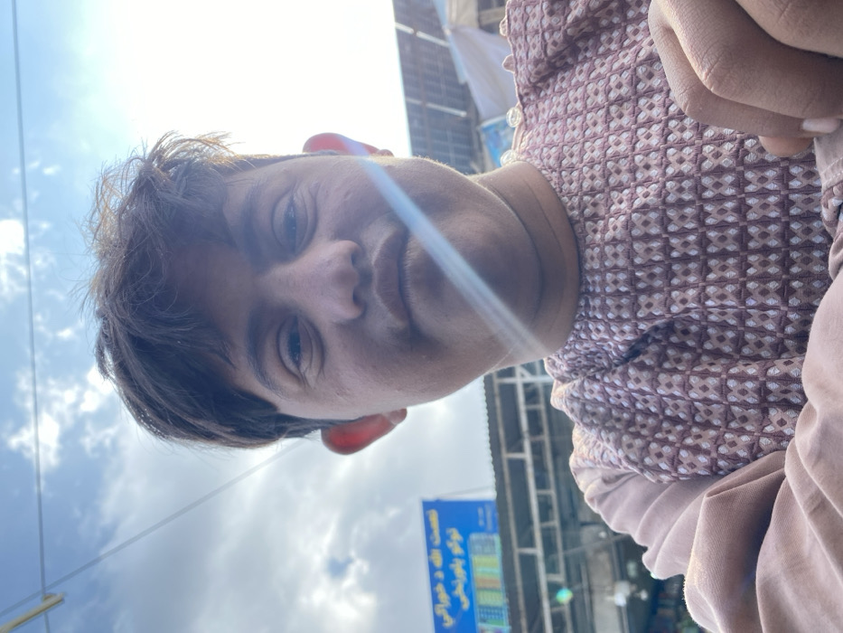This image depicts a young male captured in a low-angle close-up, likely a selfie, outdoors in a market or commercial street in Kandahar, Afghanistan. The subject, of apparent Afghan ethnicity, occupies the central and lower portions of the frame. He has dark, short, slightly disheveled hair, dark eyes, a broad nose, and full lips, which are slightly pursed. A prominent sun flare traverses diagonally across his lower face and neck. He is wearing a light brown or mauve garment with a textured, patterned weave on the upper torso, resembling small, repeating geometric shapes, with solid light-colored sleeves. His right hand is partially visible in the lower right corner.
The background reveals an urban setting under a partly cloudy sky. To the upper left, a blue signboard with white text in an Arabic script (likely Pashto or Dari) is visible, with colored rectangular panels below it. Further structural elements, possibly market stall supports or building frames, are present beneath the signboard. To the upper right, multiple solar panels are mounted on a rooftop or elevated structure. The scene is brightly illuminated by direct sunlight, casting strong light and a lens flare.