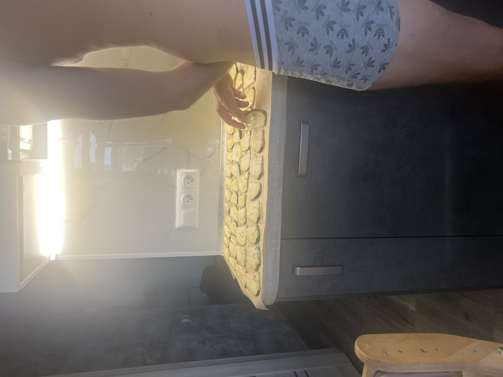 The image depicts an indoor scene, likely a kitchen, with a person in the foreground wearing grey patterned boxer briefs. The person's torso and arm are visible as they are interacting with sliced eggplant arranged on parchment paper on a countertop. The countertop appears to be dark gray, and there are kitchen cabinets below with silver handles. The background shows a white tiled backsplash illuminated by overhead lighting. Electrical outlets are visible on the wall. A wooden object, possibly a child's play structure, is partially visible on the left side of the frame. There is no indication of the location being in Volgograd, Russia.