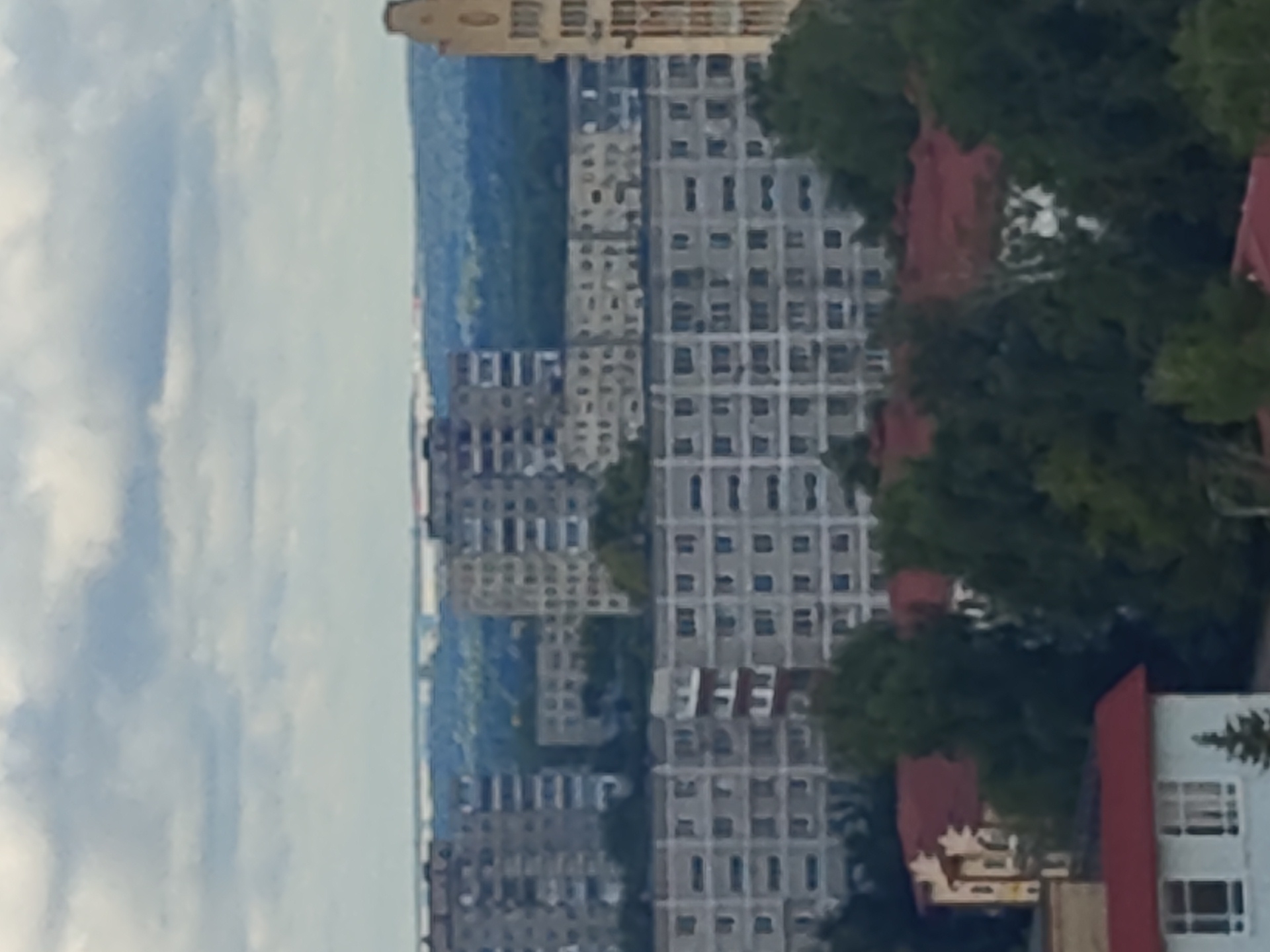 The image depicts a wide view of an urban landscape in Ramenskoye, Russia, characterized by dense residential buildings and green spaces under a cloudy sky. In the foreground, a light-colored building with a red roof is visible, featuring multiple windows and a small evergreen tree. Behind this structure, more buildings with red roofs are partially obscured by lush green trees. Further into the scene, a series of large, multi-story apartment complexes dominate the mid-ground, their grey facades and numerous windows forming a repetitive pattern. The buildings are arranged in rows, suggesting a planned residential area. In the background, a densely wooded area stretches across the horizon, interspersed with more distant buildings. The sky is filled with heavy, grey clouds, indicating an overcast or potentially rainy day. The overall impression is one of a typical Soviet-era or post-Soviet urban environment.
