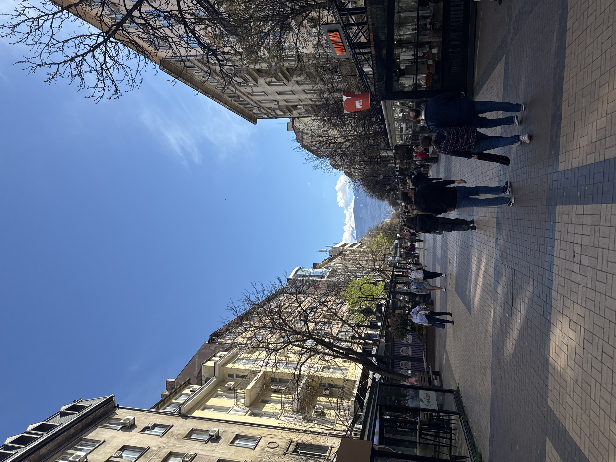The image depicts a street scene in Sofia, Bulgaria. The view is oriented along a pedestrianized boulevard lined with buildings of varying architectural styles. People are walking along the sidewalk, some in groups. The sky is blue with some clouds visible in the distance, where mountain peaks are seen. The scene is well-lit by daylight. Trees with bare branches line the boulevard. Street furniture, including benches and outdoor cafe seating, is present.