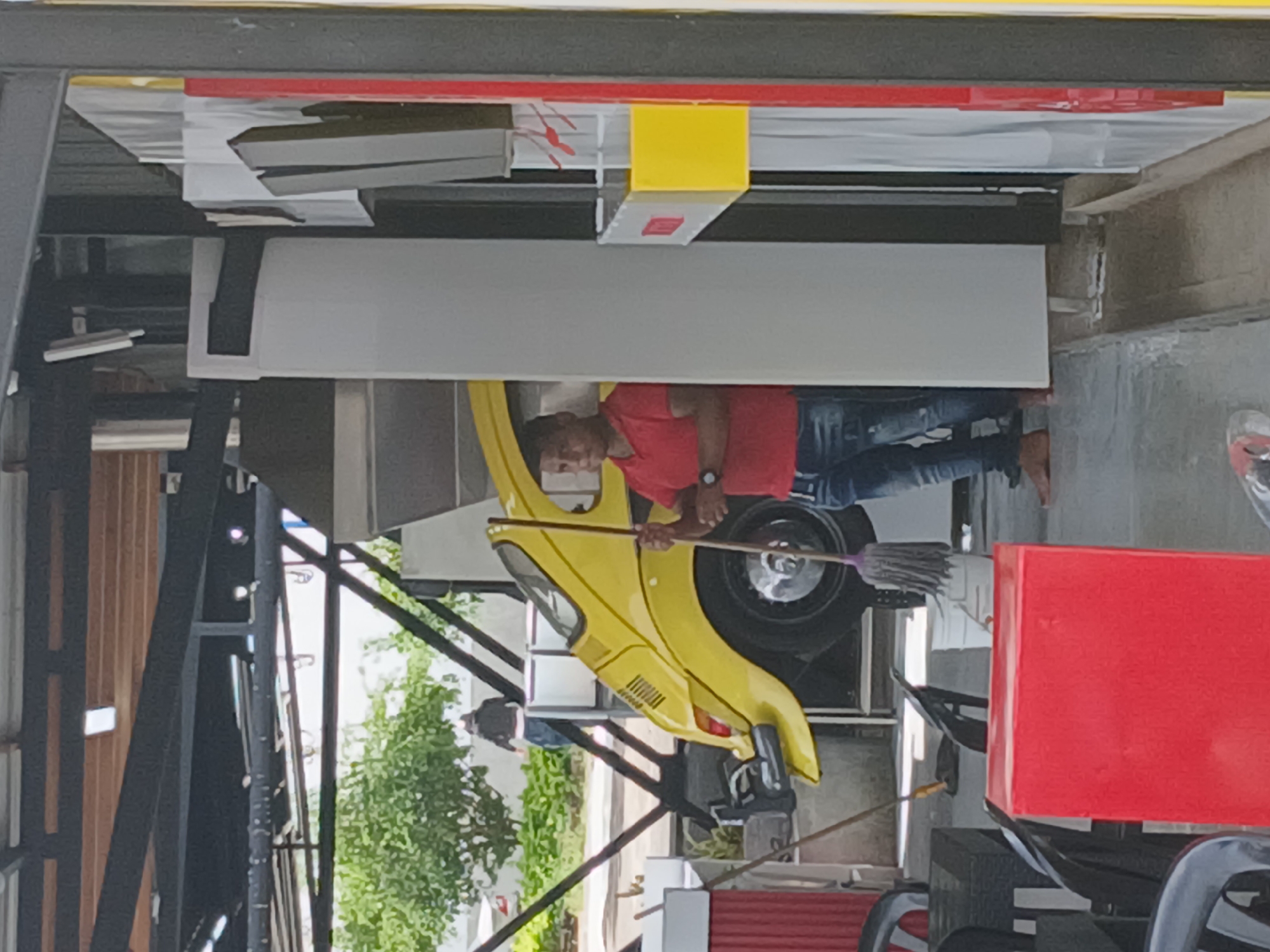 A rotated medium shot captures a man in a red shirt sweeping a yellow car in Ciudad Guayana, Venezuela. He wears a watch on his left wrist and has short dark hair. The car, possibly a modified model or decoration, sits on a wheeled stand. The environment suggests an outdoor setting, with a partial view of trees and foliage, as well as architectural elements. A red object is visible on the right side. The shot is taken from an elevated position, revealing additional structures and an enclosed area.