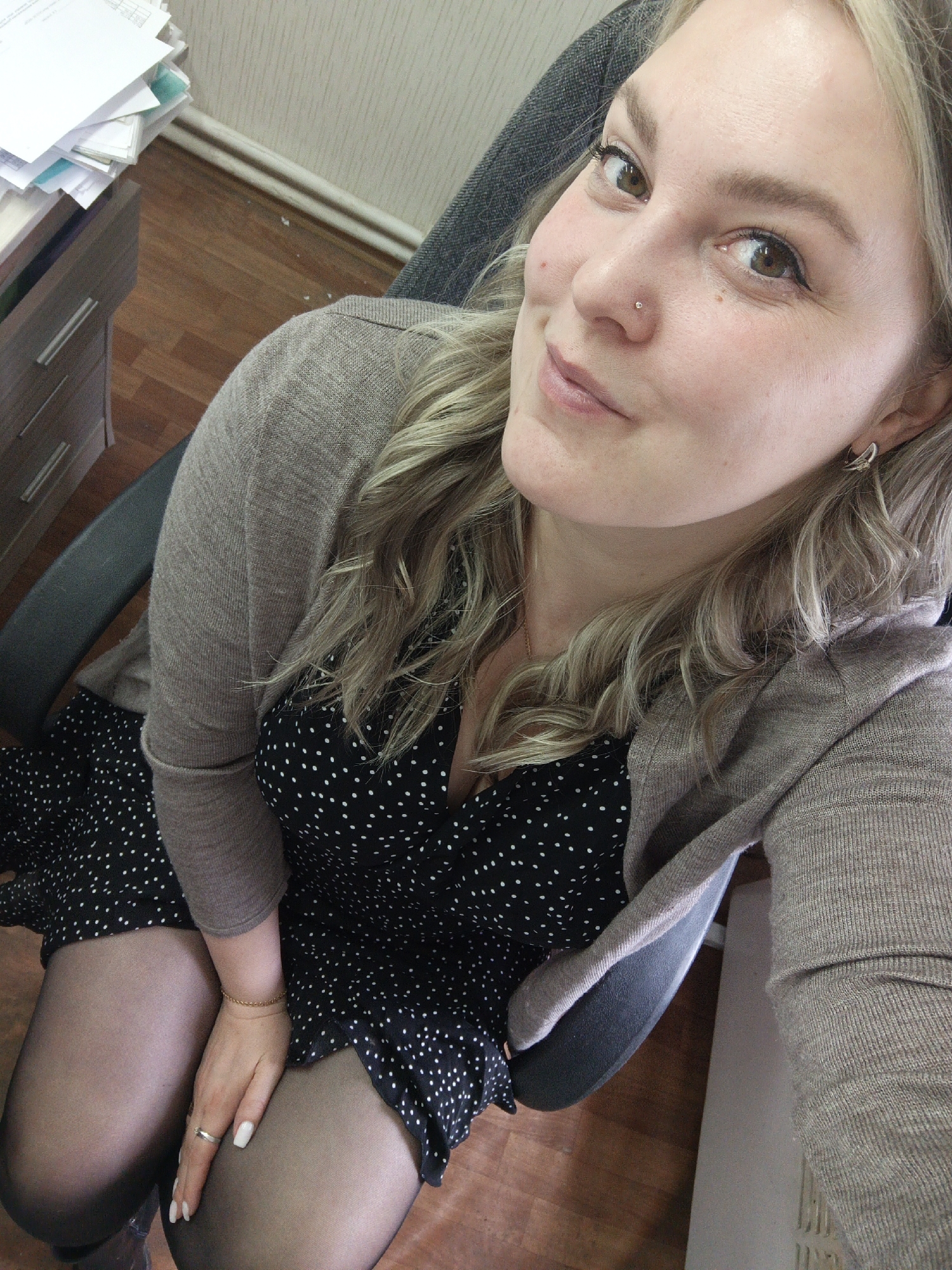 A fair-skinned woman with light blonde hair is seated in a black office chair wearing a black dress with white polka dots, black tights, and a beige cardigan. She has a nose piercing and is wearing jewelry. She is smiling and looking directly at the camera. A filing cabinet with papers on top is visible in the upper left of the frame. The floor is wooden. The location in the city of Noviy, Russia, cannot be confirmed based on the visual information provided.