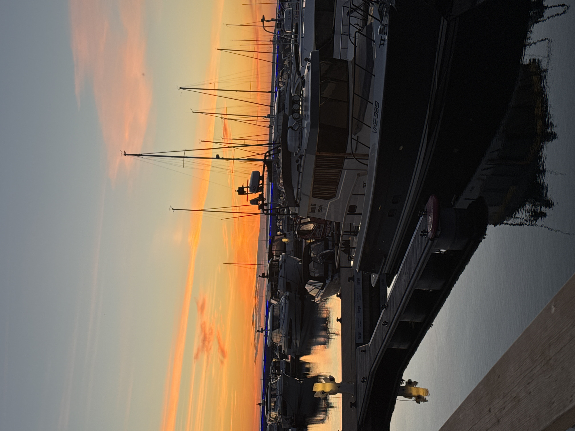 This is a photograph of a marina in Tallinn, Estonia, during sunset. The sky is filled with vibrant orange and pink hues, reflecting off the calm water. Numerous boats of various sizes are docked, with their masts forming a distinct silhouette against the colorful sky. The foreground shows a wooden pier and the calm water of the harbor. The overall scene depicts a peaceful evening at the marina.