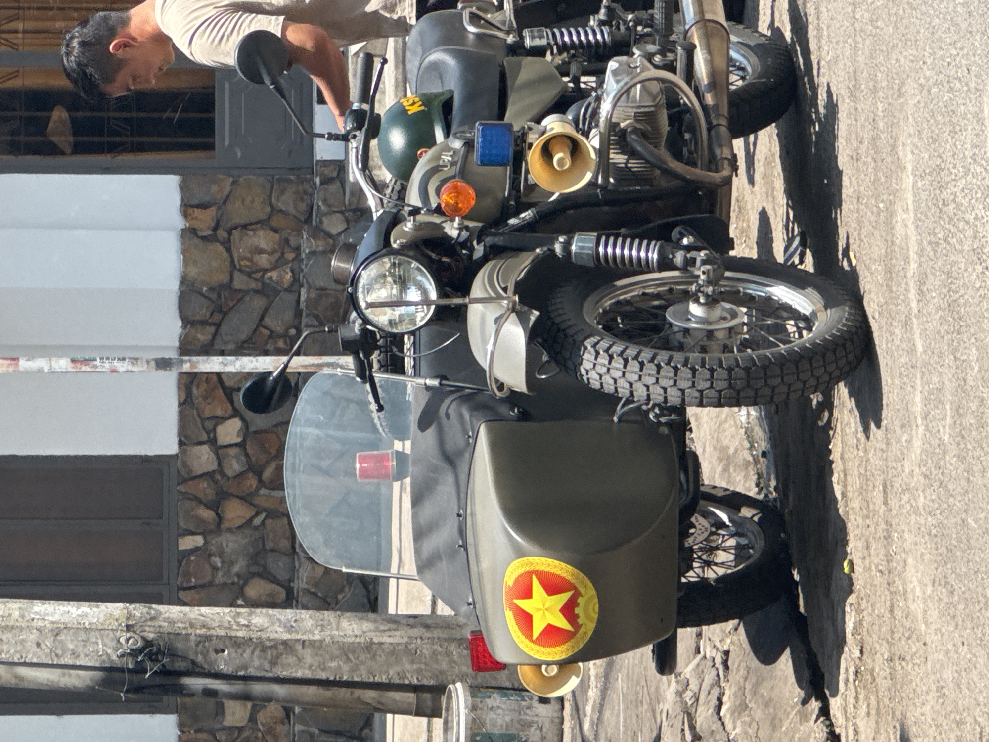 An eye-level, medium shot captures a military-style sidecar motorcycle parked on a paved street in Nha Trang, Vietnam. The motorcycle is olive drab and features a sidecar on the left, adorned with a red and yellow star emblem. A red flashing light is visible on the windshield of the sidecar. The motorcycle also has a blue light and a megaphone-like speaker mounted on its front. A man in a light-colored t-shirt is standing to the right of the motorcycle, appearing to be engaged with it. The background shows a building with stone and white facade, and a utility pole on the left. The street is cracked and appears well-trafficked.