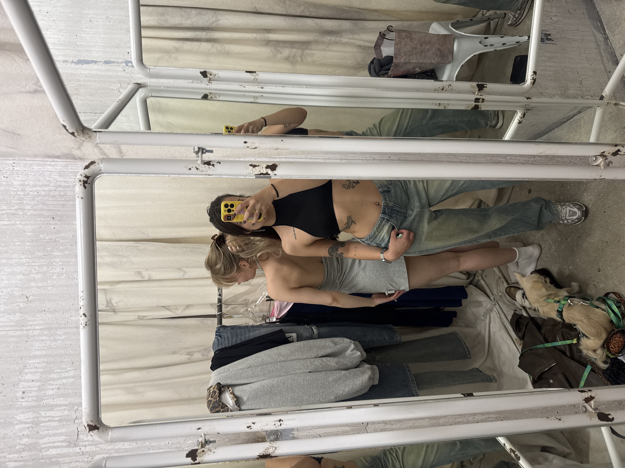 A mirror selfie captures two young women and their dog within what appears to be a rustic or industrial-style indoor space, possibly a fitting room or a market stall in Moscow, Russia.
The primary subject, a woman with dark hair, is visible in the reflection of a large, multi-panel mirror. She wears a black crop top and light blue, distressed jeans, showcasing tattoos on her arm and stomach, and a belly button piercing. She holds a bright yellow smartphone with a dark camera module and a finger ring holder, actively taking the mirror selfie.
Standing beside her, also reflected in the mirror, is a woman with blonde hair tied back with a hair clip. She is wearing a light grey strapless dress and looks slightly downwards or away from the camera. Her hands are near her waist.
Below the women, stacked on a surface or shelf, are various folded clothes, including dark blue jeans, grey sweatpants, and a black garment. On the concrete floor, next to the blonde woman's legs, a small, light-colored dog with a green and patterned harness rests next to a brown leather jacket and a patterned tote bag. A single white shoe is also visible nearby.
The setting is characterized by a multi-panel mirror structure with a white, heavily rusted metal frame. The walls behind the mirror appear to be painted concrete or stucco, showing signs of wear with peeling or chipped paint. The overall impression is casual and unpretentious. There is no clearly legible text visible within the image.