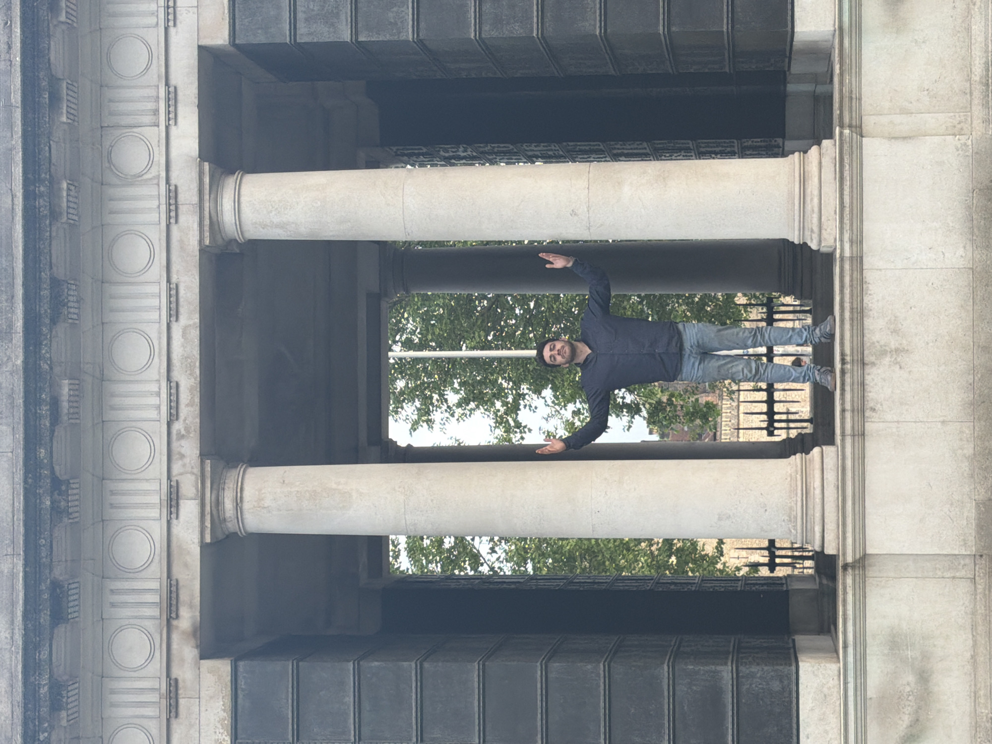 A man stands between two rows of stone columns in a colonnade. He wears a dark blue button-down shirt, light blue jeans, and gray sneakers. His arms are outstretched to the sides, his palms facing forward. Behind him, through the colonnade, trees and building facades are visible. The architectural style appears classical, with decorative details on the structure surrounding the columns. This is likely the National Portrait Gallery in the city of London, United Kingdom. The image is presented on its side with the top of the photo pointing towards the left.