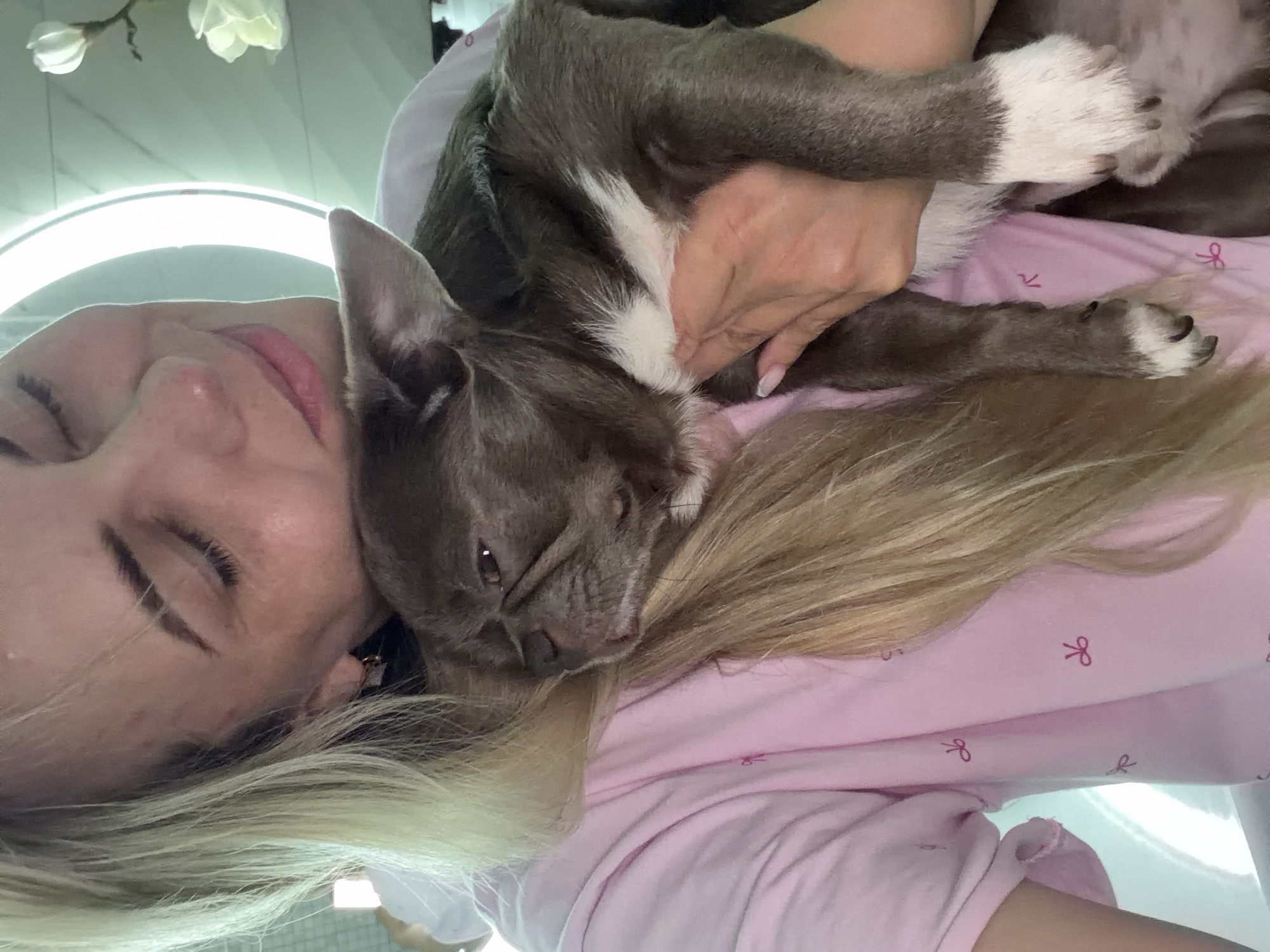 The image displays a close-up, intimate moment between a woman and a small dog, both appearing to be resting or sleeping indoors. The woman, positioned on her side, has light blonde hair and closed or partially closed eyes, with a relaxed expression. She is wearing a light pink top patterned with small red bows. A small, grey-brown dog, possibly a French Bulldog, with distinctive white markings on its chest and paws, is snuggled against her head and hair, its own eyes closed in apparent comfort. One of the woman's hands, with neatly manicured nails, gently supports or caresses the dog. The background features a prominent bright circular light source, likely a ring light, and some artificial white flowers, indicating an indoor setting. The overall atmosphere conveys peace, affection, and comfortable companionship. No visible text is present in the image. The location context provided is Votkinsk, Russia.