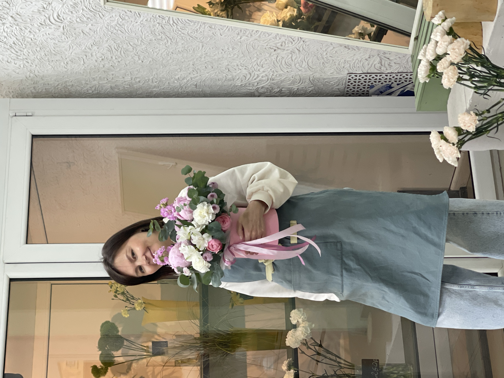 A woman with dark hair is holding a bouquet of flowers in a pink box with pink ribbons tied around it. She is wearing a white sweater and a grey apron. The scene appears to be indoors, possibly in a flower shop, as there are more flowers and plant displays visible through the glass window behind her. The location is Unknown, Unknown. The ceiling has a patterned texture.