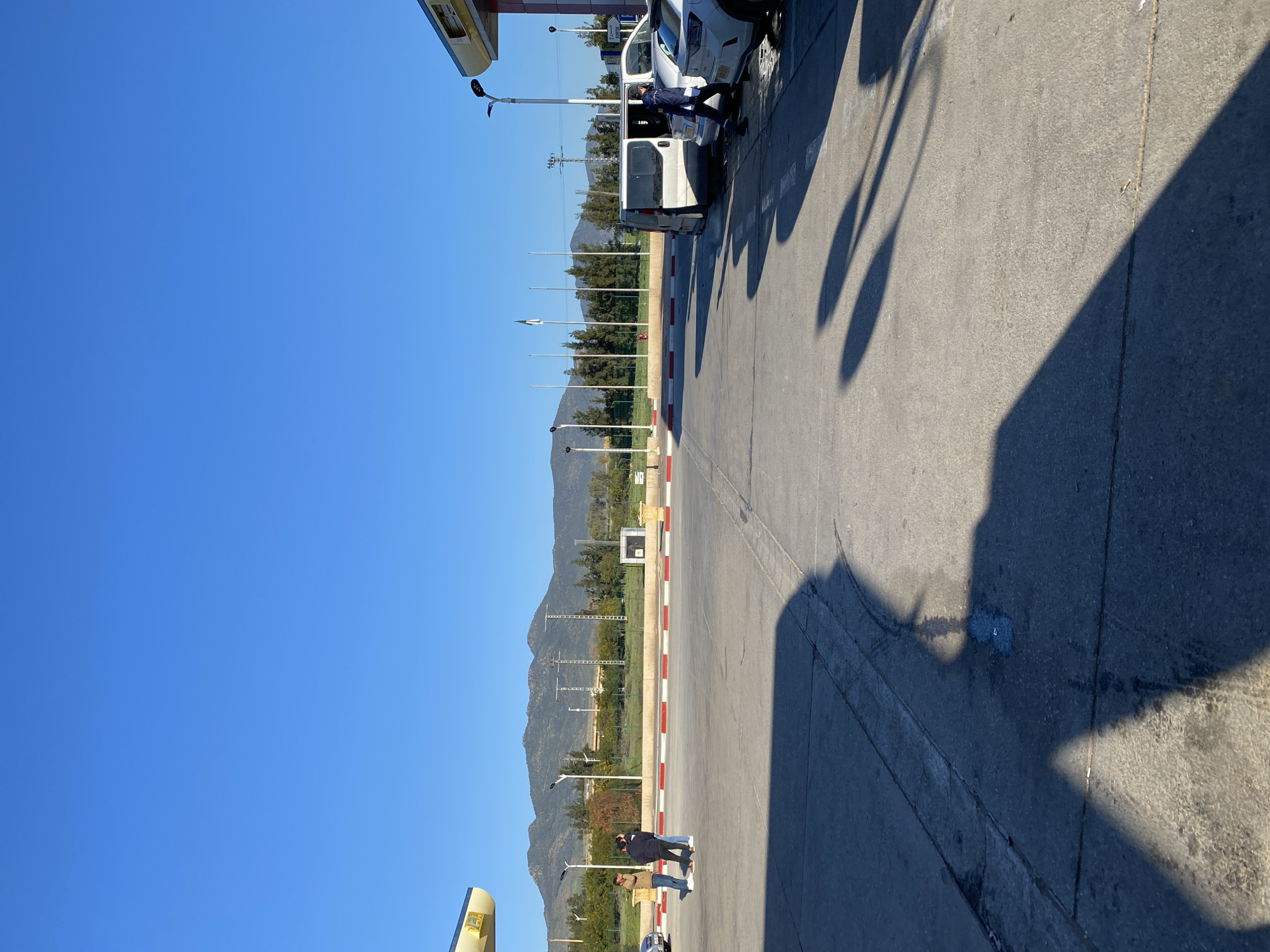 A daytime shot in Djelida, Algeria, shows a roadside scene with mountains in the background. A white van is parked near a building with a yellow overhang. A person in a dark jacket is interacting with the van, while another person stands nearby. Streetlights cast shadows on the asphalt surface. A red and white striped barrier is visible along the edge of the road. The sky is clear and blue.