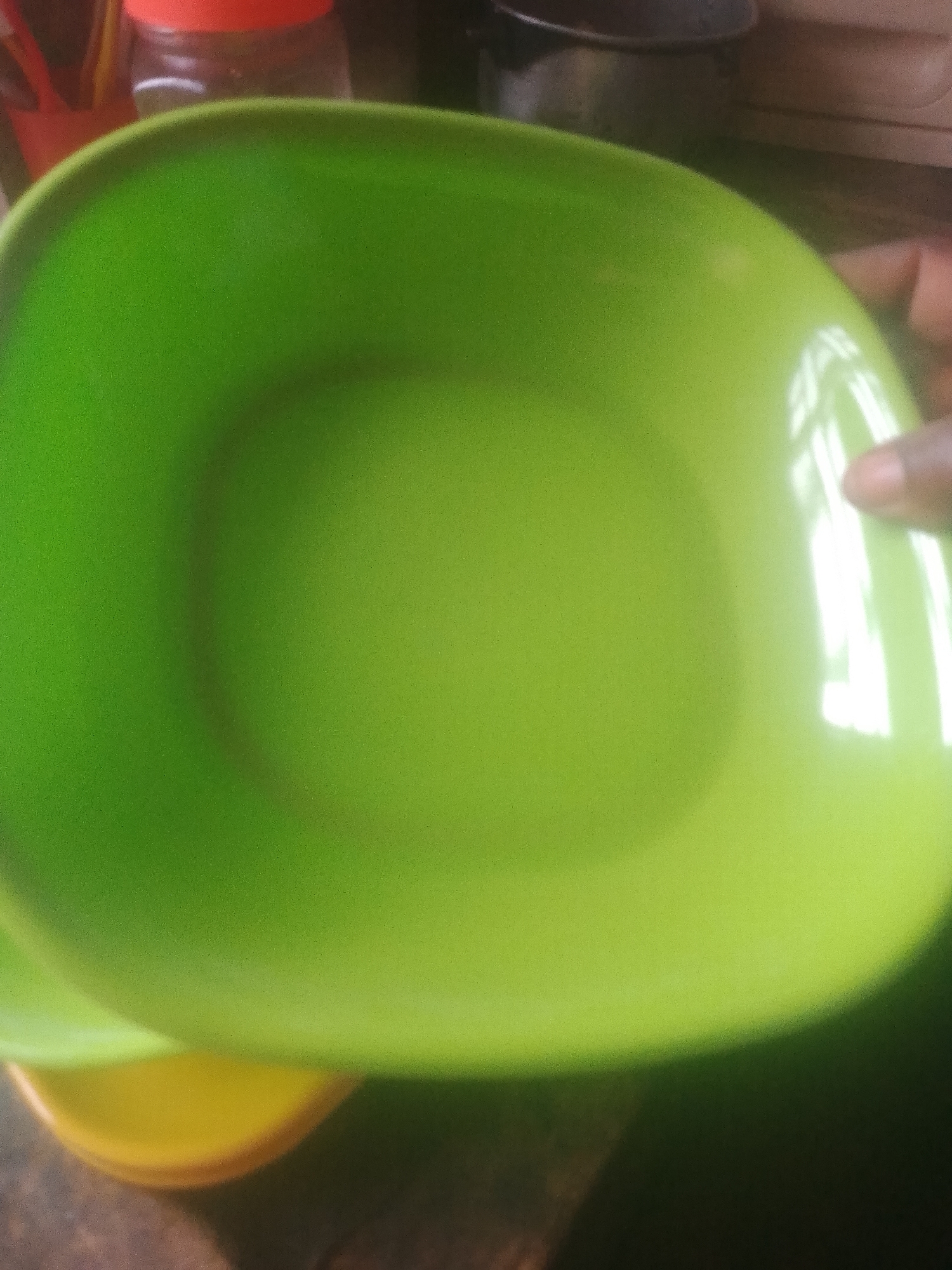 A bright green plastic plate is centrally placed in the foreground, angled from the lower left to the upper right of the frame. The plate has a slightly raised rim and a smooth, reflective surface that catches the light, creating bright white highlights, particularly on the right side. Beneath the green plate, partially visible at the bottom, is a yellow plate or bowl. In the blurred background, there are hints of other objects, including what appears to be a red-capped container on the left and dark kitchenware on the upper right. A person's hand, with a light skin tone, is visible on the far right edge of the image, appearing to hold or interact with the green plate. The overall lighting is somewhat dim, with the primary light source creating strong reflections on the plate. The scene is set indoors, likely in a kitchen environment, and is not specific to Jauro Yinu, Nigeria.