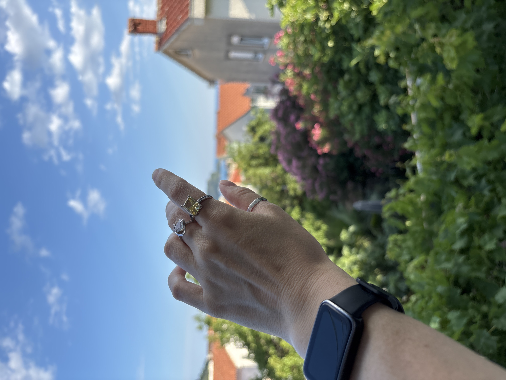 A human hand with three rings on its fingers and a smartwatch on its wrist is extended towards the viewer, pointing at the sky. The background depicts a sunny day in Budva, Montenegro, with a clear blue sky filled with white clouds, and a cluster of houses with red-tiled roofs nestled amongst lush green trees and flowering bushes. The scene captures a moment of contemplation or observation against the backdrop of a picturesque Mediterranean town.