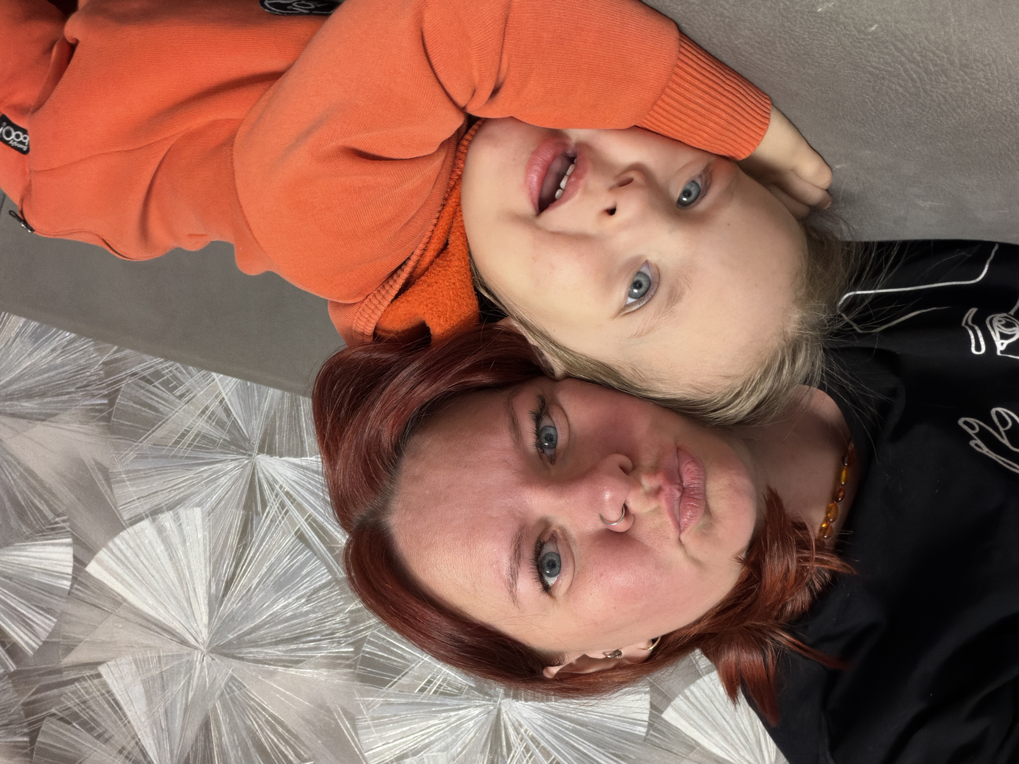 The image depicts two individuals: an adult female and a young child. They are positioned close together, their heads adjacent, and appear to be lying down. The child is wearing an orange sweatshirt and has blonde hair. The adult female has reddish-brown hair and is wearing a black top. The background consists of a gray surface and a white and silver patterned surface. There is no specific information in the image to suggest the location is in Chaikovsky, Russia.