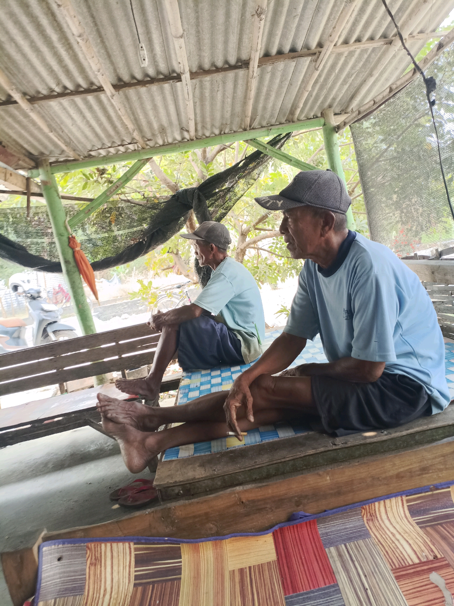 The image depicts two elderly Asian men sitting on a wooden bench under a corrugated metal roof supported by green painted metal poles. The man in the foreground, facing right, wears a light blue t-shirt, black shorts, and a grey baseball cap. His legs are crossed, and he holds a cigarette in his right hand, which rests on his knee. The second man, in the background and facing left, wears a similar light blue t-shirt and dark shorts, also with a cap. Both men appear to be relaxed. Behind them, a motorbike and a bicycle are visible, suggesting an outdoor, possibly rural or semi-urban setting. The environment includes trees and foliage peeking through the roof structure. The scene is likely in Rancang Kencono, Indonesia, as per the provided context, suggesting a local Indonesian atmosphere.