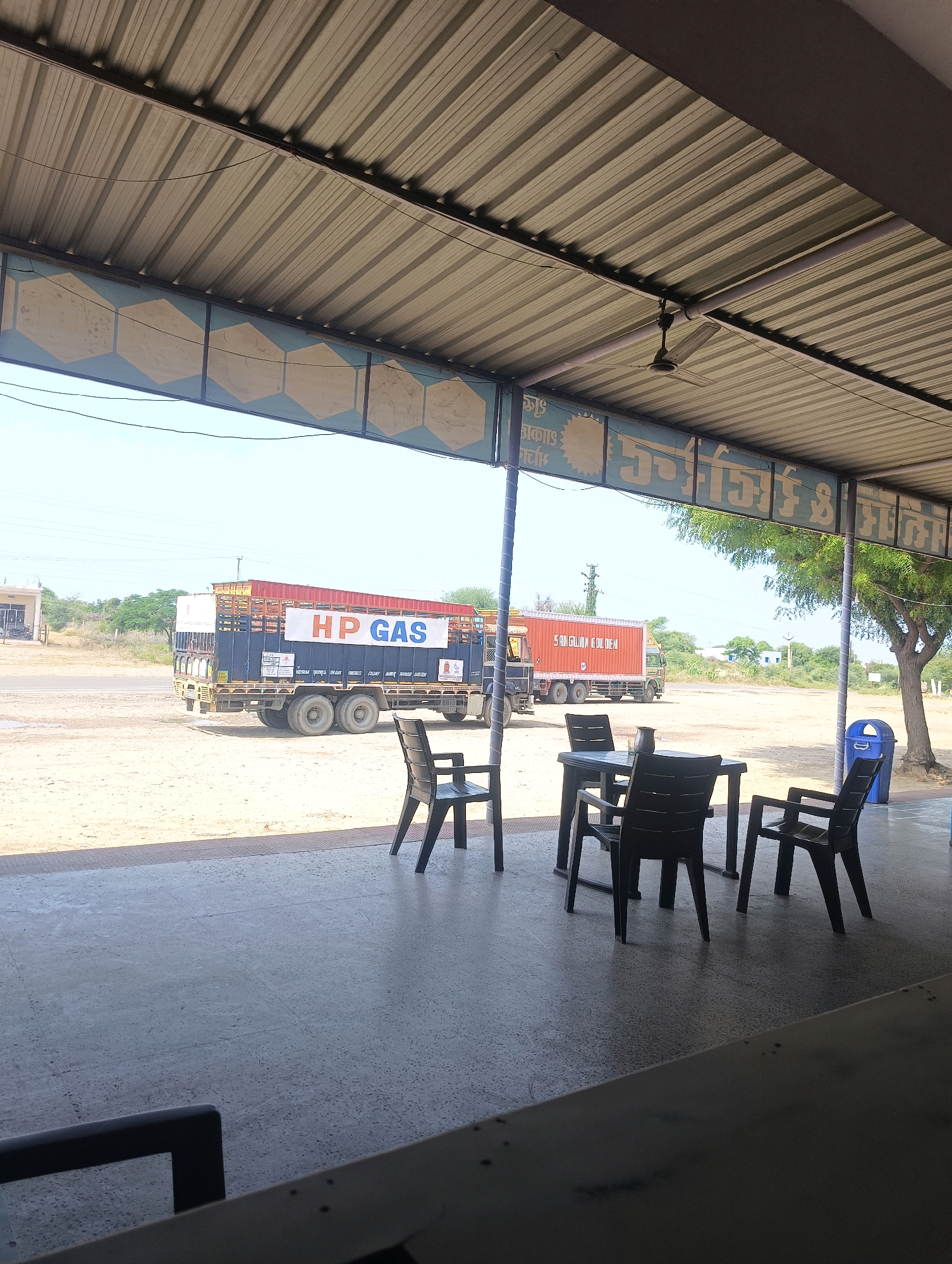 A wide shot captures an outdoor seating area at a highway rest stop in Chhapri Bari, India. The foreground is dominated by a grey concrete floor. A table with four dark plastic chairs is set up, with a small metal jug on the table. In the mid-ground, a large truck with "HP GAS" written on its side is parked on a dusty surface. The truck is dark blue with a red cargo area. Beyond the truck, a few trees and buildings are visible under a bright, clear sky. The scene is partially shaded by a corrugated metal roof supported by pillars. A blue trash can stands to the right of the seating area.