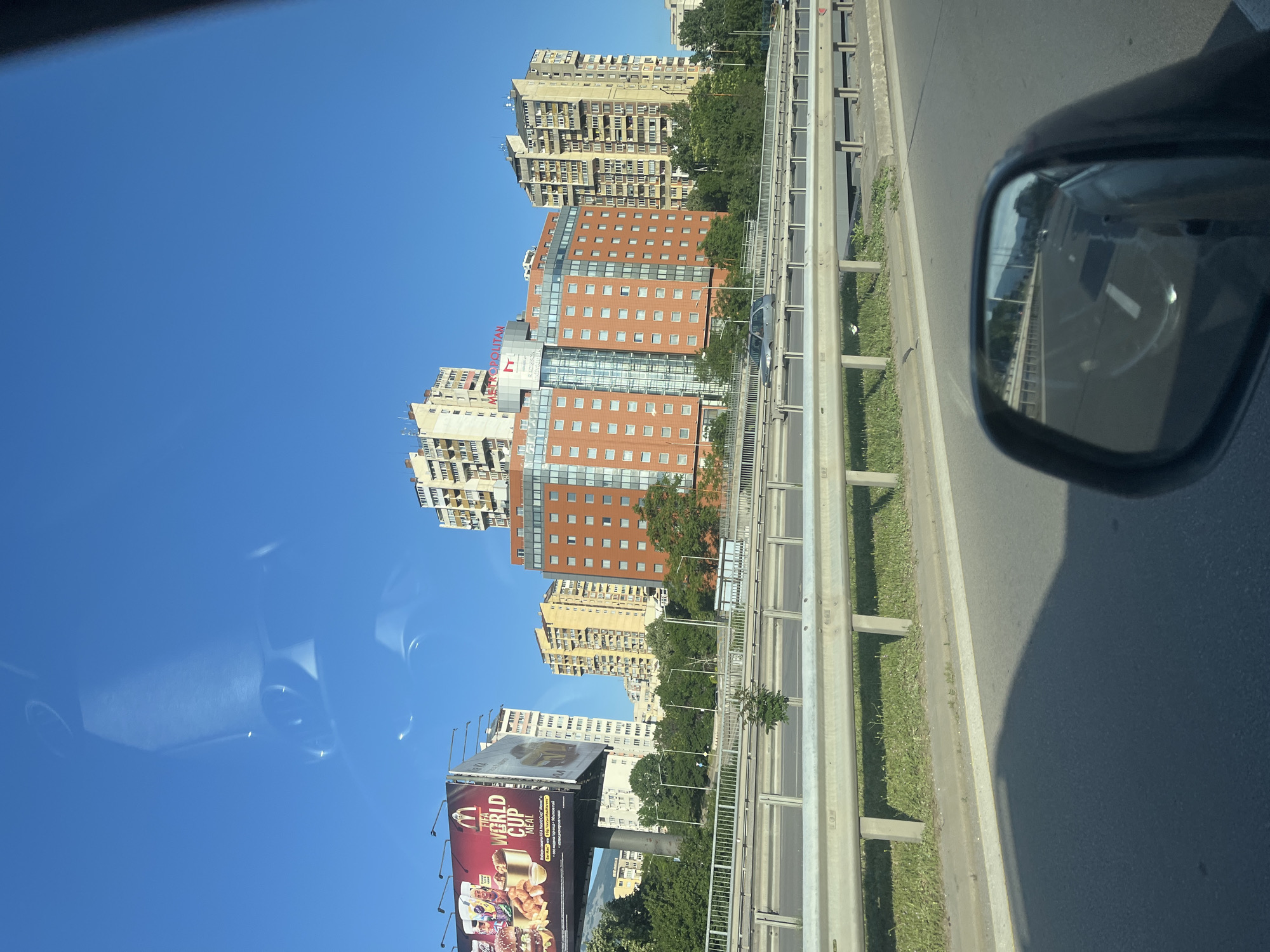 The image captures a view from a car traveling on a highway in a city. The sky is clear and blue. On the left side of the highway, a large billboard advertising McDonald's "FIFA World Cup Meal" is visible, along with several tall buildings. In the center and right of the frame, a modern hotel with an orange facade, branded "METROPOLITAN," stands prominently, surrounded by other apartment buildings. A silver car is driving in the opposite direction on the highway. The foreground shows the road surface and a metal guardrail separating the lanes, with a strip of grass and some trees beyond. The side mirror of the car reflects the highway extending into the distance, with other vehicles and buildings also visible in the reflection.