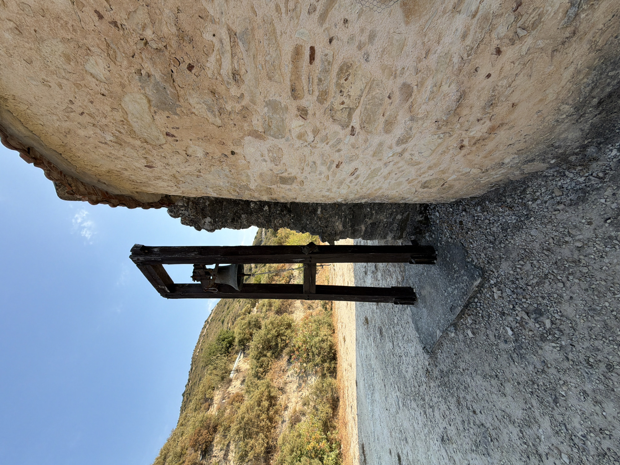 The photograph shows a weathered bell on a wooden frame situated next to a stone building in the village of Agios Amvrosios, Limassol, Cyprus. The bell, made of metal, hangs from the wooden structure, which appears aged and dark. The building to the right has a rounded facade constructed from rough-hewn stones of varying sizes and colors, coated with a pale plaster. A section of the roof, tiled with terracotta, is visible at the top. The ground in front of the bell and building is covered with gravel and dirt. Behind the bell, a dry, scrub-covered hillside slopes upward, dotted with sparse vegetation and trees under a clear blue sky. No people are present in the image.