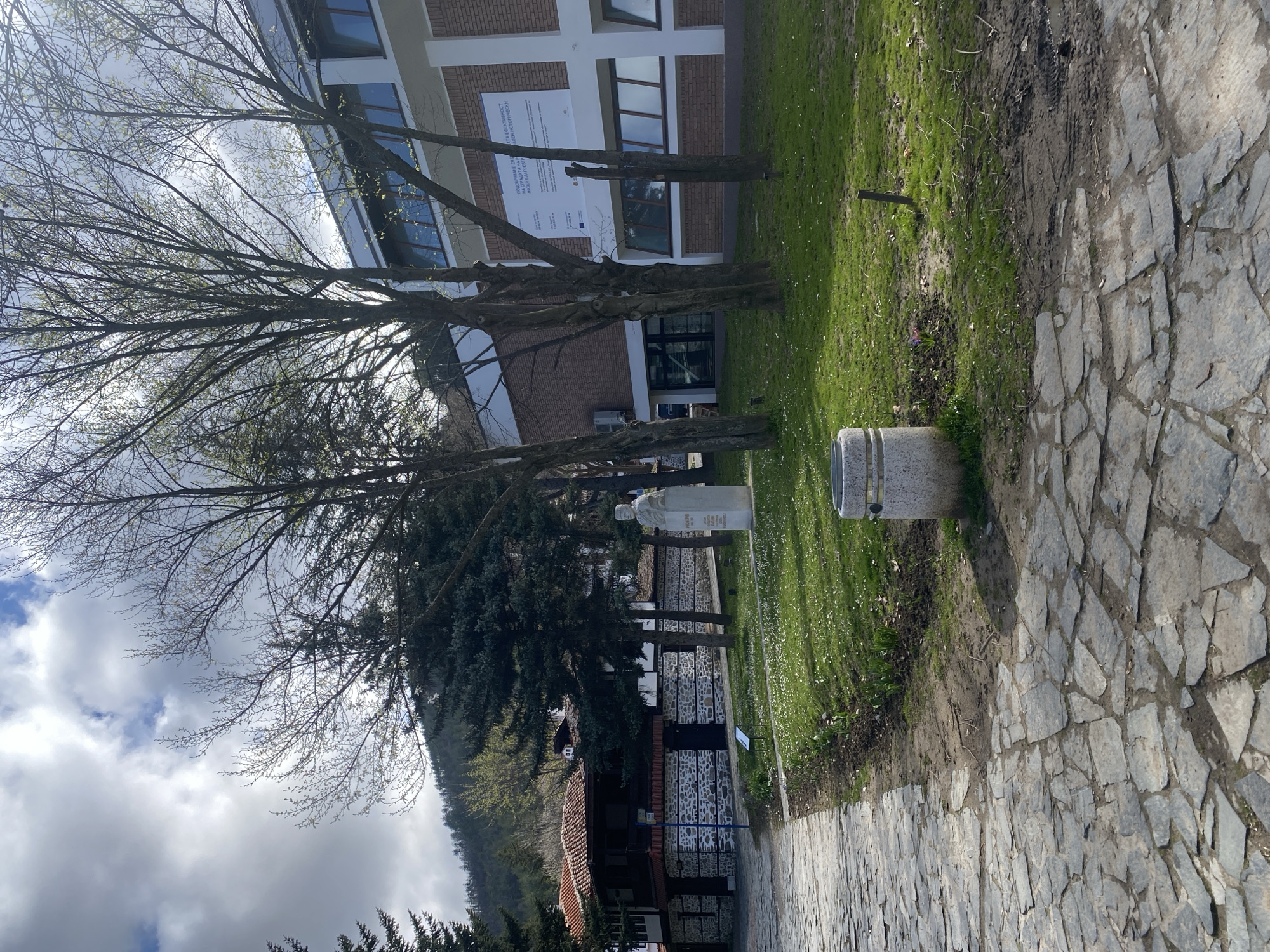 The image depicts a scene in Blagoevgrad, Bulgaria. A stone pathway occupies the foreground, leading to a grassy area with trees, a statue, and a stone trash receptacle. Beyond the grassy area is a building with a brick facade and several windows. A large sign is visible on the building's exterior. Behind the building, a hillside is partially obscured by trees. The sky is cloudy.