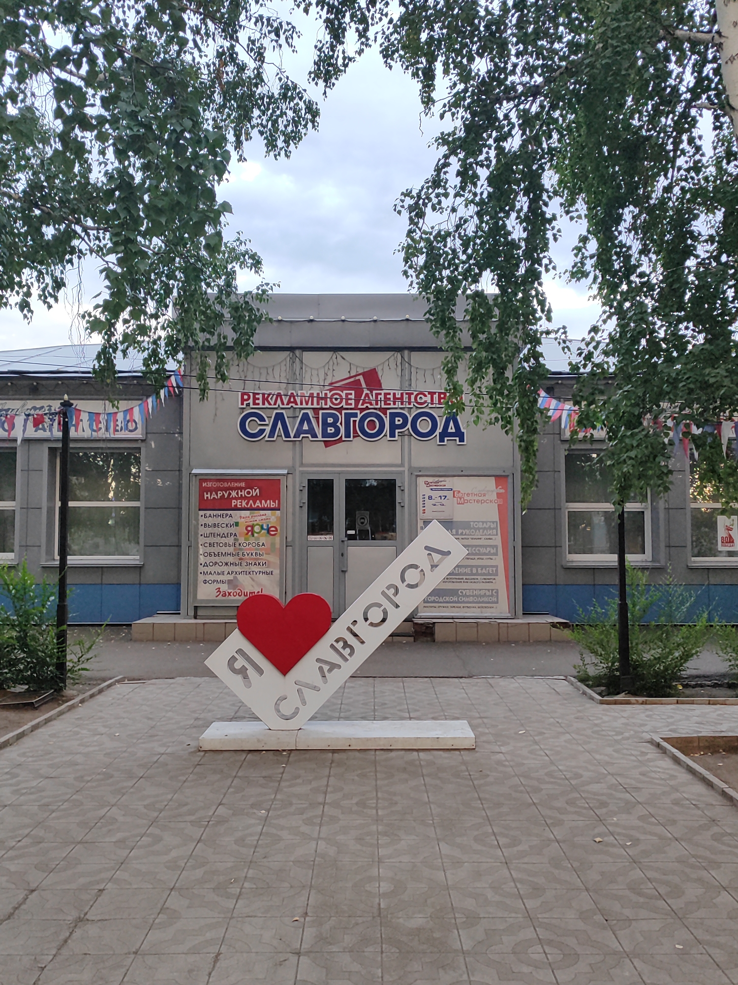This media file captures the exterior of a building in Slavgorod, Russia, likely a business. The building's facade is a light gray with blue accents near the bottom. Above the entrance, prominent Russian text reads "РЕКЛАМНОЕ АГЕНТСТВО СЛАВГОРОД" which translates to "ADVERTISING AGENCY SLAVGOROD."
In front of the building, a prominent white sculpture in the shape of an angled "I" followed by a heart and the word "СЛАВГОРОД" (Slavgorod) is displayed. This suggests a sense of local pride or a tourist attraction. The sculpture is made of white material with cut-out letters.
A red heart is positioned on the "I" part of the sculpture, indicating affection or love for the city. The building features multiple windows and two glass doors for entry. To the left of the main entrance, a smaller red sign lists services offered by the agency, including banners, signs, light boxes, volumetric letters, road signs, and small architectural forms, with the word "Заходите!" (Come in!) at the bottom. To the right, another sign indicates "Багетная Мастерская" (Framing Workshop) and mentions "ТОВАРЫ ДЛЯ РУКОДЕЛИЯ" (Craft Supplies) and "СУВЕНИРЫ С ГОРОДСКОЙ СИМВОЛИКОЙ" (Souvenirs with city symbols).
The scene is framed by the branches of leafy trees, suggesting it's a daytime shot with overcast or cloudy skies. The ground in front of the building is paved with square tiles. Small, colorful Russian flags are strung along the building, adding a festive touch. The overall impression is a commercial establishment with a welcoming and community-oriented message.