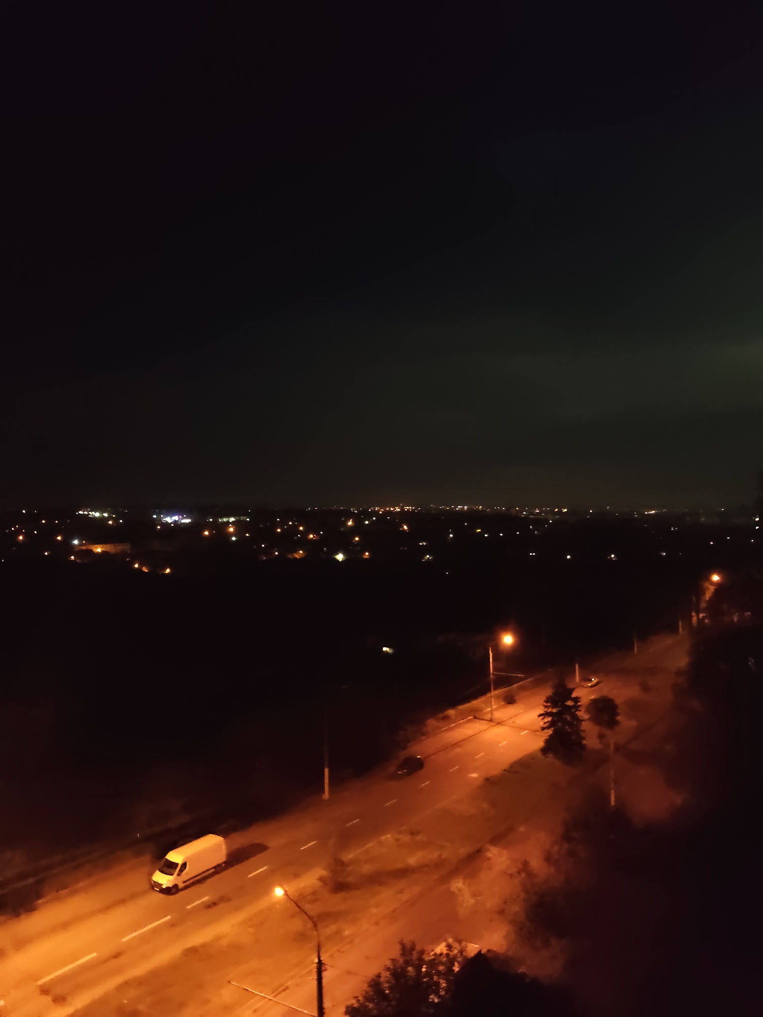 This is an aerial, nighttime photograph overlooking a city that is likely Zaporizhzhia, Ukraine, due to the distinct orange glow of streetlights typical of the region and the general urban landscape. In the foreground, a white van is traveling on a two-lane asphalt road, which is illuminated by the orange glow of streetlights. The road has white lane markings. To the right of the road, there are trees and more streetlights. In the middle ground, the road curves and other vehicles, including a dark car, are visible. Further in the distance, a sprawling cityscape is illuminated by numerous lights, indicating a densely populated area. The sky above is dark with some faint cloud cover. The overall impression is of a city at night, with active transportation and urban infrastructure.