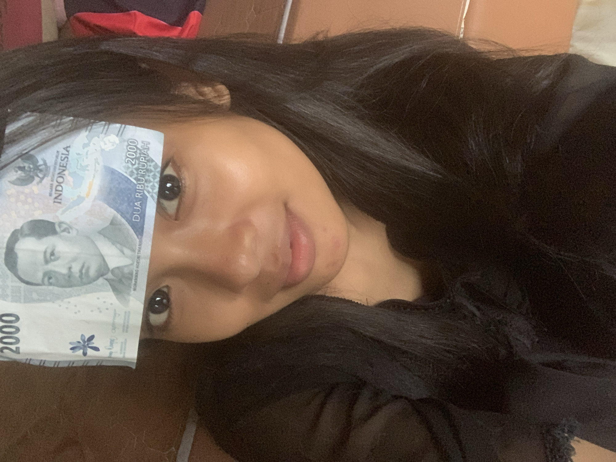 A young person with long, dark hair is seen lying down in a close-up, angled shot. Her face, primarily her right eye, part of her left eye, and her mouth, are visible. She gazes directly at the camera with a soft, slightly smiling expression. An Indonesian 2000 Rupiah banknote is placed across her forehead, covering part of her left eye.
The banknote prominently displays "INDONESIA", "2000", "DUA RIBU RUPIAH", and "NEGARA KESATUAN REPUBLIK INDONESIA". A portrait of Mohammad Hoesni Thamrin and the Garuda Pancasila emblem are also clearly visible on the currency, which appears to be from the 2016 series. The person is wearing dark clothing, likely black. The setting appears to be indoors, possibly a bedroom or living area, with a muted background. The image is well-lit, showing details of the person's skin and hair. The overall impression is one of a casual, relaxed pose, possibly in Sungai Meriam, Indonesia.
