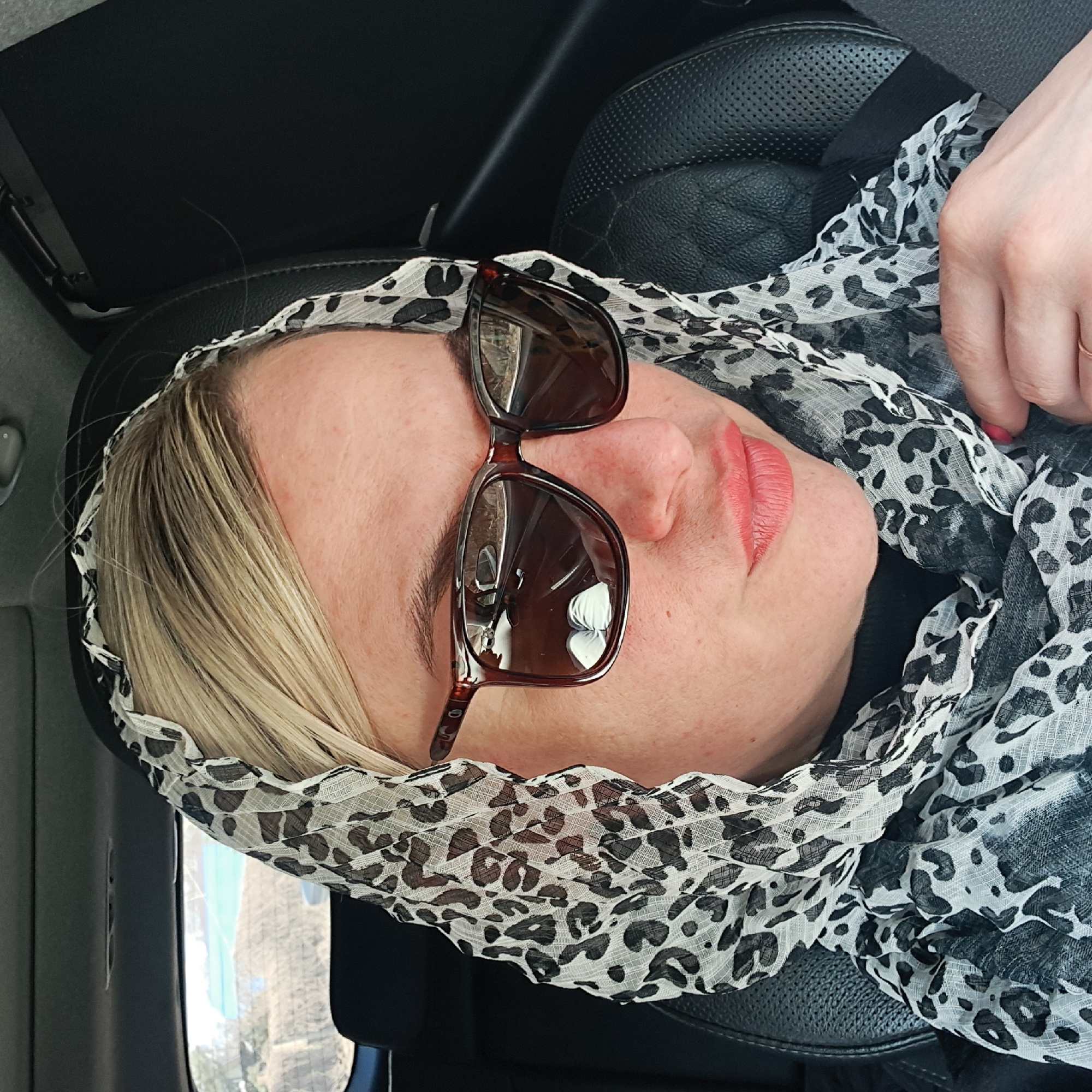 A woman with fair skin and blonde hair sits in what appears to be the driver's seat of a car. She is wearing a black and white leopard-print scarf covering her head, and brown-framed sunglasses are perched on her nose. Her lips are painted with a coral or light red lipstick. Part of her hand is visible near her chest, with red nail polish on her fingernails. The interior of the car includes black leather or pleather seats. The background visible through the car window suggests an urban or suburban environment, but specific landmarks are not discernible enough to confirm the location of Elektrougli, Russia.