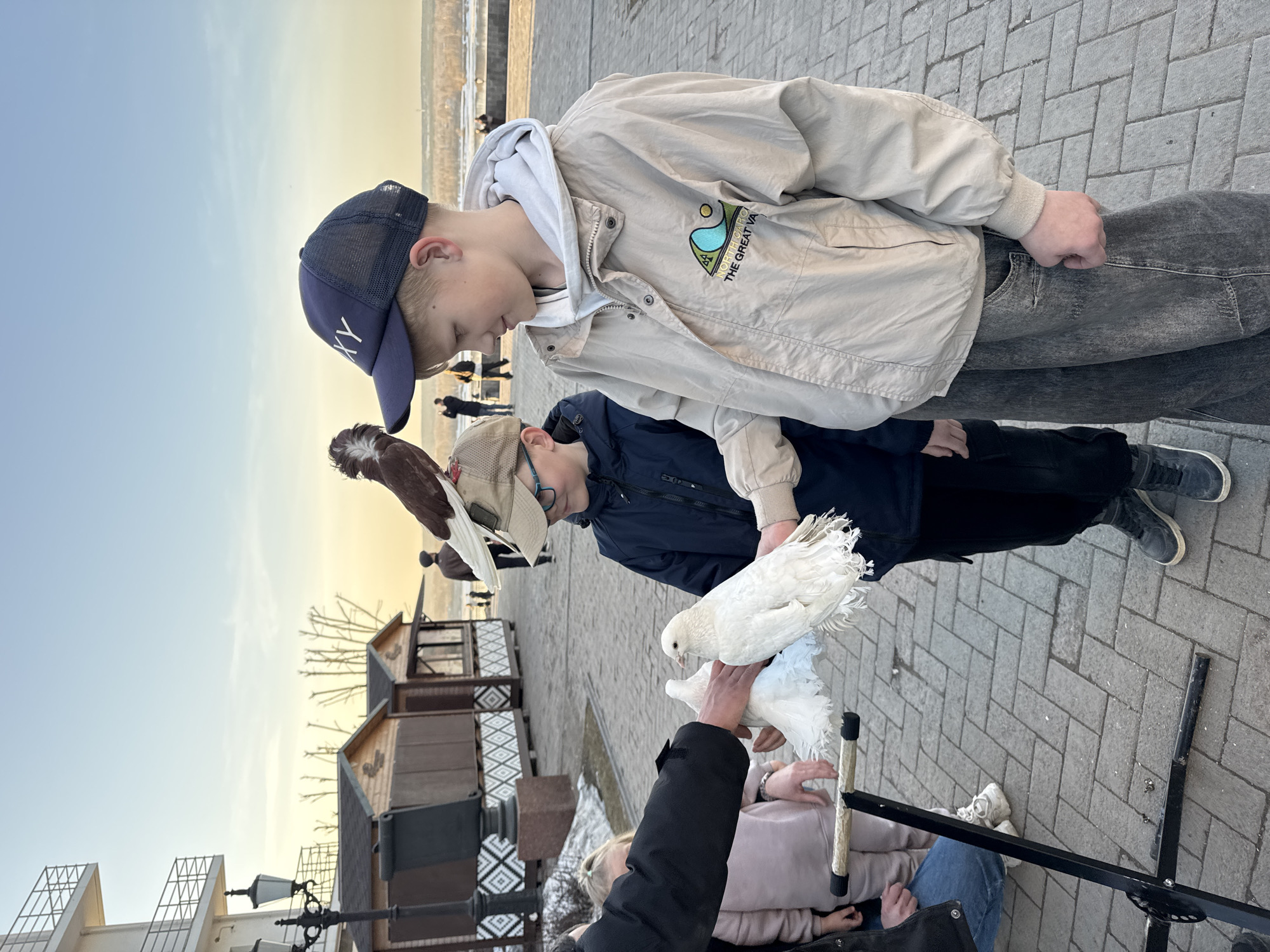 Two boys stand on a paved walkway, looking at white doves being held by someone whose lower half is visible. The older boy wears a light jacket, gray hoodie, and a navy cap. The younger boy wears a navy jacket and a tan cap. A brown and white bird is perched on the younger boy's cap. The background shows a waterfront, small wooden structures, and other people in the distance. The location is Perm, Russia, near the waterfront.