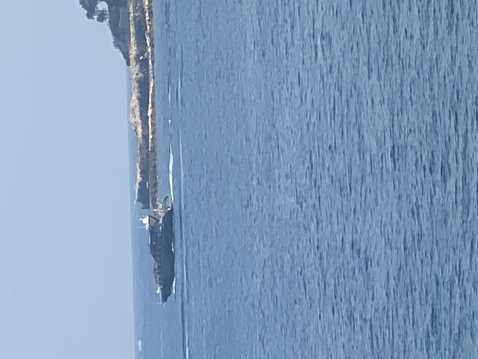 A dark-colored motor yacht cruises across a bright blue ocean, leaving a white wake behind it. The yacht is positioned on the left side of the image, moving from left to right. In the background, a rocky coastline with sparse vegetation is visible on the right. The sky is a clear, pale blue. The scene is consistent with the coastline of Antibes, France, possibly near the Cap d'Antibes area, given the rocky outcrops and the presence of a yacht in the Mediterranean Sea. There are no people clearly visible on the yacht or in the surrounding environment. The image appears to be taken from a distance, likely from another vessel or the shore.