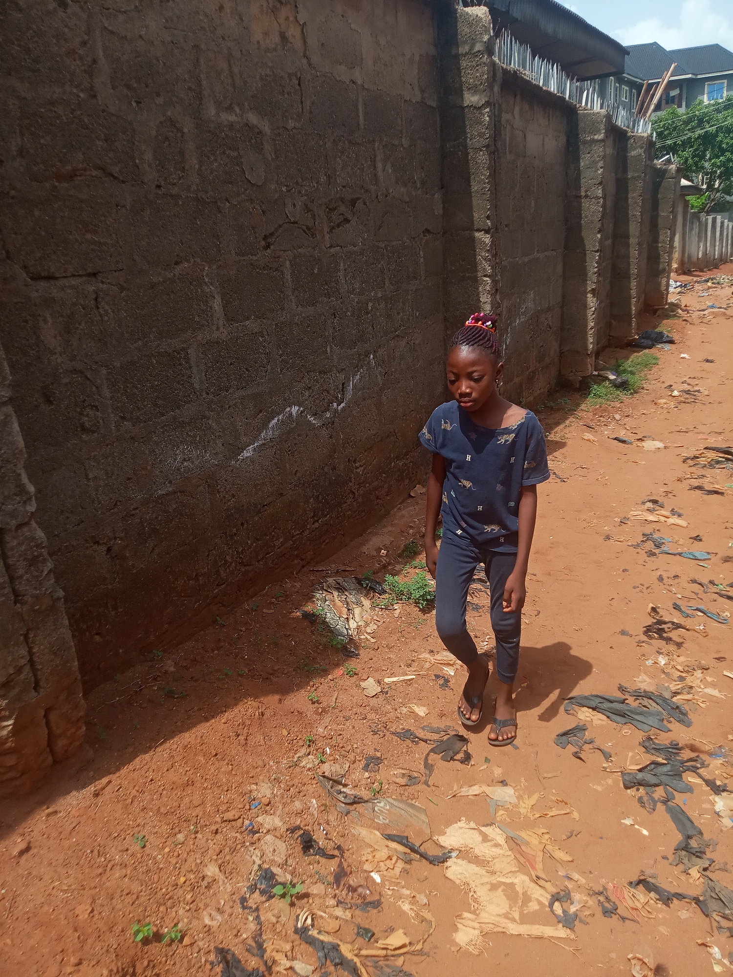 A young girl walks along a reddish-brown dirt path next to a tall, weathered concrete block wall under bright daylight. The girl, with braided hair adorned with colorful red and purple bands, wears a dark blue t-shirt featuring light-colored patterns, possibly depicting animal prints and the letter 'H', along with dark blue trousers and dark flip-flops. Her gaze is directed downwards, focused on the path ahead.
The ground is unpaved, made of reddish-brown earth, and is heavily littered with various debris, including black and light-colored plastic bags, pieces of fabric, and small patches of green weeds. Strong shadows from the girl and the adjacent wall fall across the path, indicating a sunny, likely mid-day or early afternoon, with a clear sky visible in the background.
The concrete wall, constructed from large blocks, appears aged and somewhat rough, with visible cracks and some white markings. The top of the wall is fitted with a corrugated metal overhang and sharp metal anti-climb spikes. In the far background, behind the wall, parts of a building with a dark roof and a green tree are discernible. The scene depicts an outdoor setting in what appears to be a residential or semi-urban area, consistent with the location context of Onitsha, Nigeria. The girl's expression is neutral, showing no strong emotions.