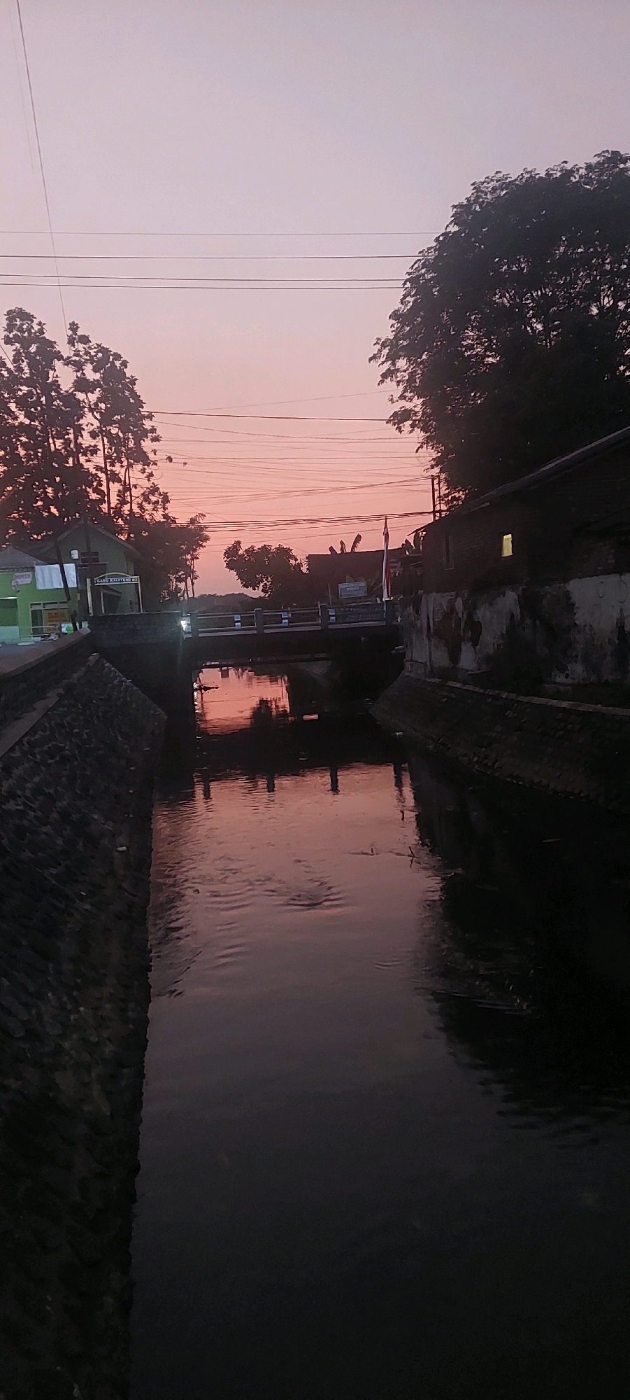 The image depicts a canal in Lumajang, Indonesia, at dusk. The sky displays a soft gradient of pink and purple hues, characteristic of a sunset. A concrete bridge spans the canal, with streetlights illuminating its walkway. On the left bank, stone walls are visible, supporting a sidewalk and a building with a green facade. On the right bank, a darker, more rustic building with visible brickwork and a lit window stands. Trees with dense foliage frame the scene on both sides, silhouetted against the colorful sky. The water in the canal is dark and reflects the sky and surrounding structures, creating a serene and tranquil atmosphere. No people are visible in the image. The overall scene suggests a quiet evening in a residential or semi-urban area of Lumajang.