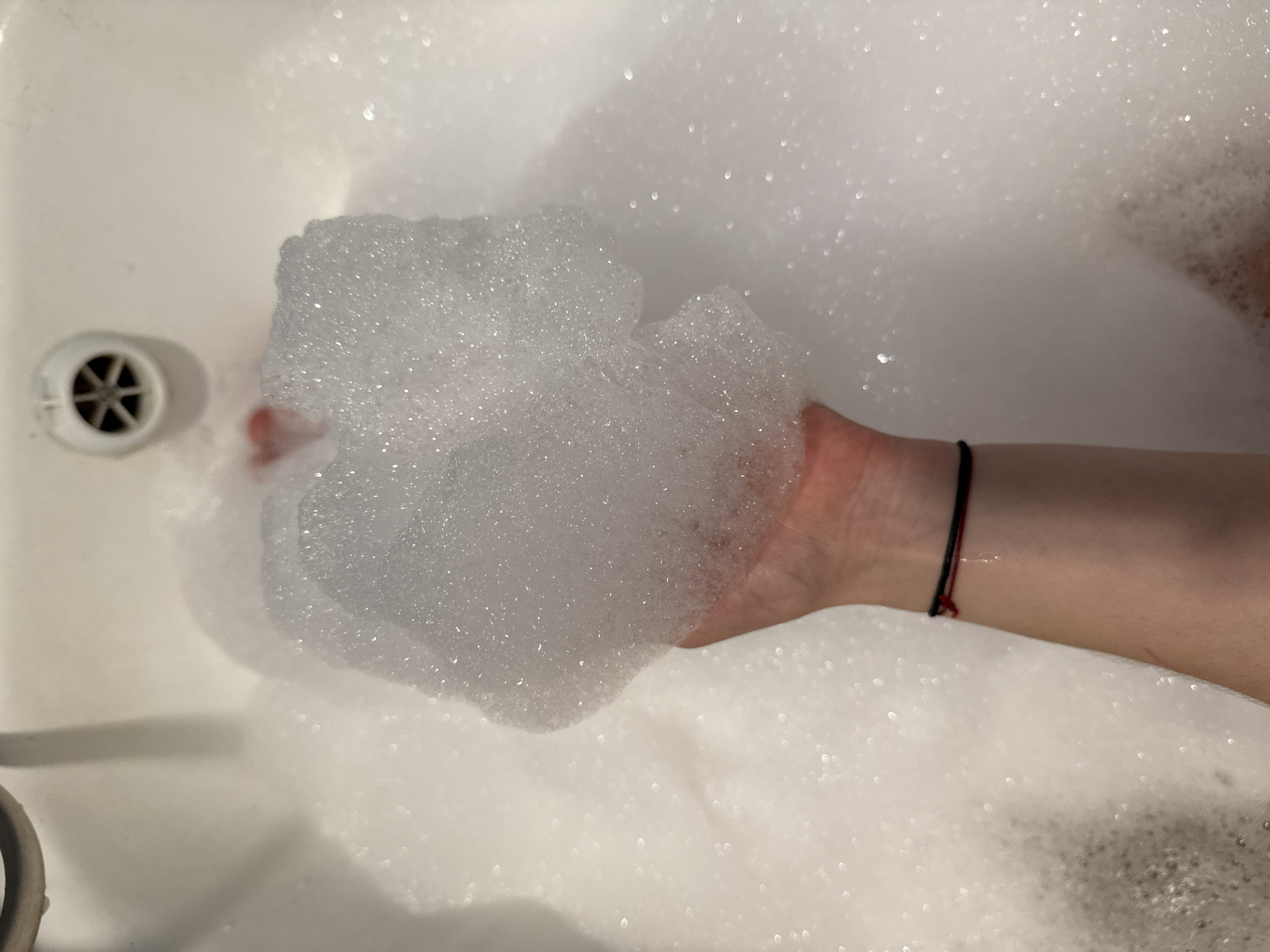 A hand holds a clump of white bath bubbles in a white bathtub. The drain is visible on the left side of the tub. A black and red cord bracelet is on the wrist of the arm holding the bubbles. The bathtub is filled with bubbles. The location of the city of Chaikovsky, Russia, is not determinable from the image.