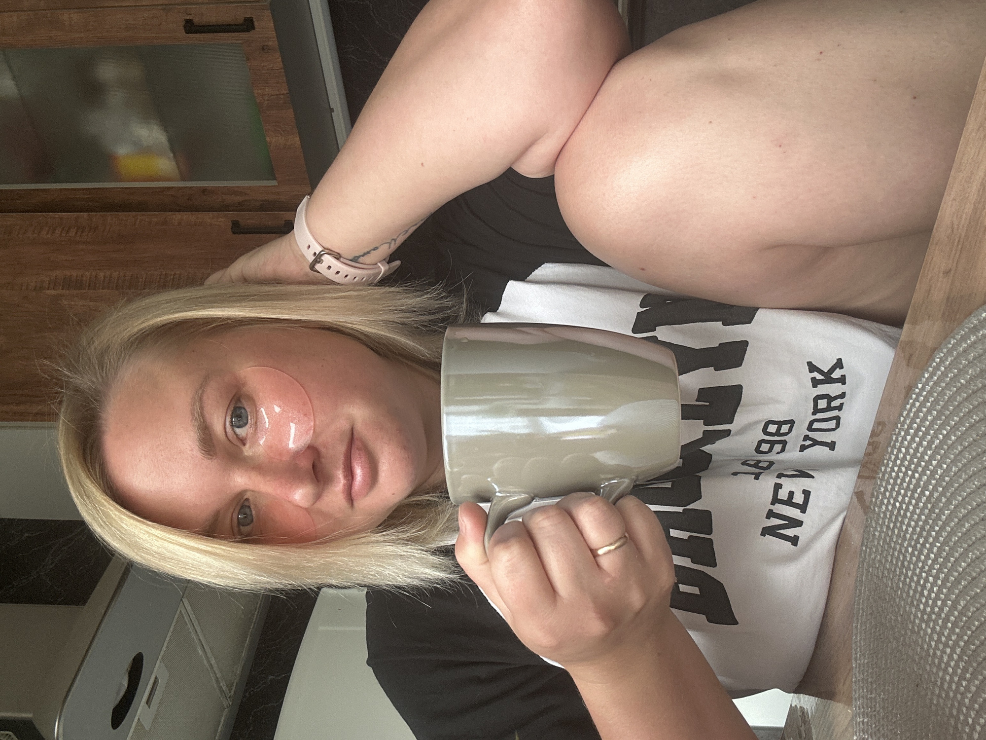 A medium shot shows a woman sitting at a table with a mug of coffee in her hand and eye patches on her face. She has blonde hair and is wearing a black and white t-shirt with the words "BROOKLYN 1898 NEW YORK" printed on it. A pink smartwatch is visible on her wrist. The background shows kitchen cabinets, a stove hood, and a countertop. The scene appears to be indoors, in a domestic setting. There is no information in the image that can be used to determine its location in Chaikovsky, Russia.