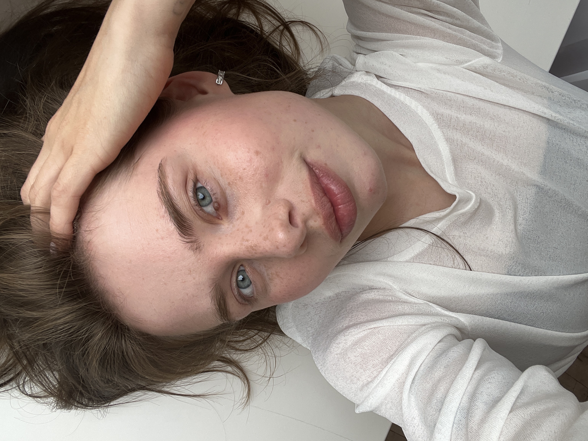 A person with fair skin and freckles, wearing a sheer white top, is lying on a white surface. Their face is turned towards the camera, with blue eyes and full lips. One hand is resting on their face. The background is white. The image does not contain enough information to determine its location in the city of Chaikovsky, Russia.