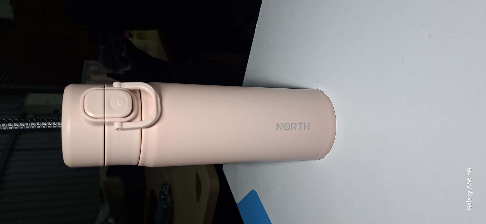 A light pink vacuum flask with a silver lid mechanism and a silver ring handle is positioned upright on a white surface. The flask has the word "NORTH" printed vertically in silver text on its body. The background is dark and out of focus, showing what appears to be a metal corrugated wall and some indistinct objects. A metallic spring is visible extending from the top of the flask. The bottom left corner of the image shows a corner of the frame with text that reads "Galaxy A56 5G". The scene does not provide any information related to the city of Kommunarka, Russia.