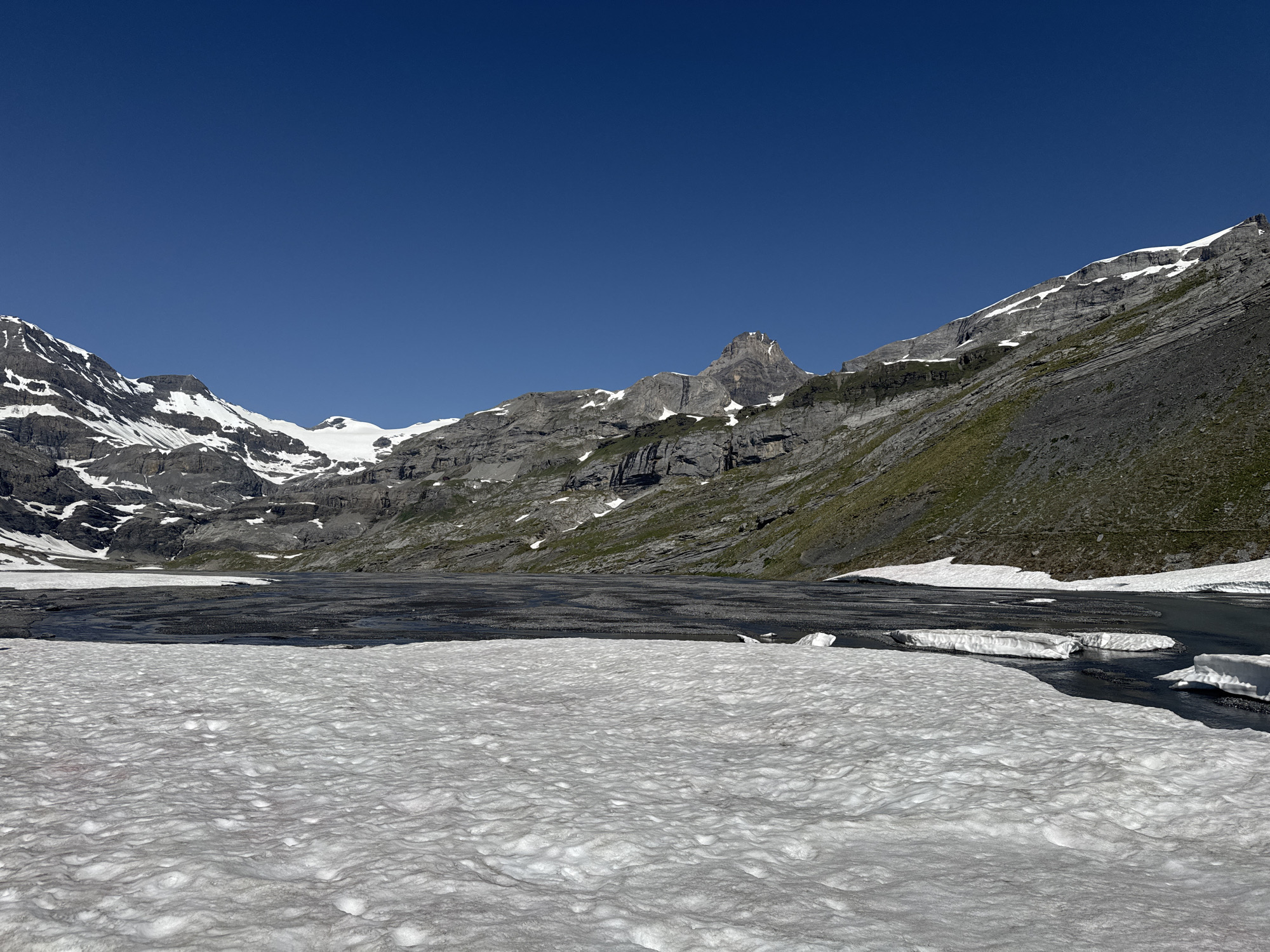 The image depicts a mountain landscape in Leukerbad, Switzerland, under a clear blue sky. In the foreground, a large expanse of textured snow covers the ground, appearing somewhat melted and uneven. Beyond the snow, a dark, rocky surface with patches of water is visible, suggesting a partially frozen lake or riverbed. Further in the background, imposing mountains rise, characterized by jagged gray rock faces interspersed with patches of white snow. The slopes of the mountains are partially covered with green vegetation. The overall scene conveys a sense of rugged, alpine wilderness during a period of transition, possibly late spring or early summer, when snow is melting. There are no people or animals visible in the image.