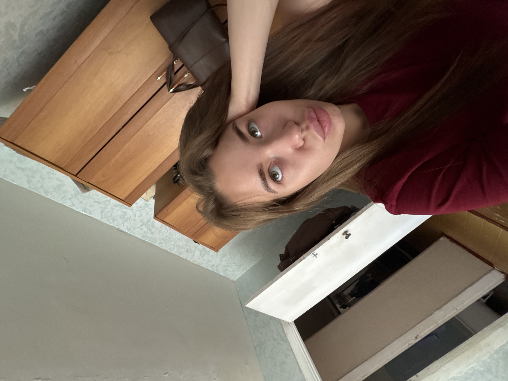 A young woman with long brown hair, wearing a red shirt, is taking a selfie in a room. She is leaning her head on her hand and looking upwards with her lips pursed. In the background, there is a wooden wardrobe, a brown handbag, and an open door revealing another room. The room appears to be a domestic interior, possibly a bedroom or living area, located in Chaikovsky, Russia. The lighting suggests it is daytime, and the overall atmosphere is casual.