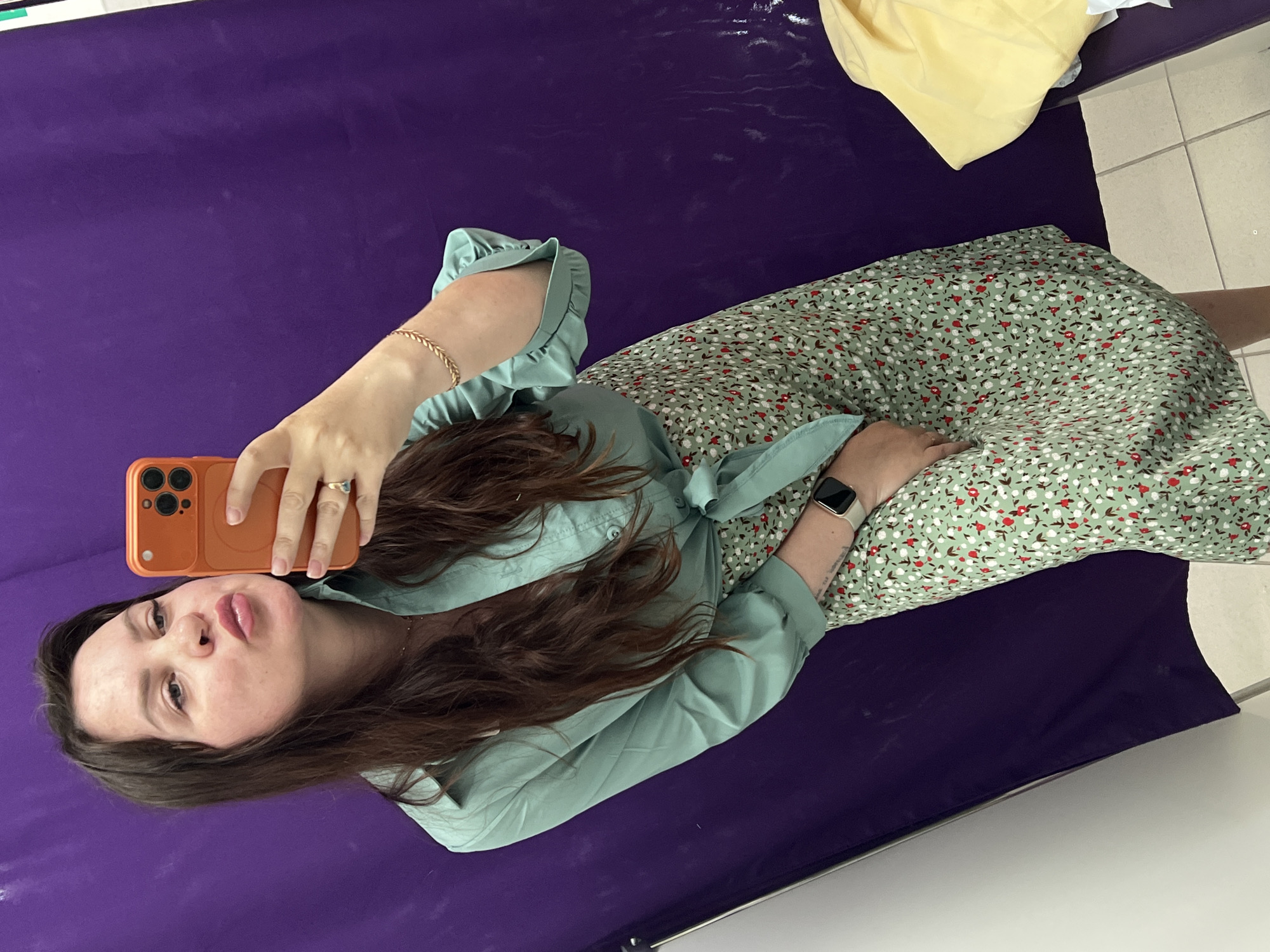 A woman is taking a selfie. She is wearing a green dress with a floral pattern and a light green blouse. She is holding an orange smartphone in her left hand. She is wearing a gold bracelet on her left wrist and a ring on her left index finger. She is wearing a smartwatch on her right wrist. The background is a purple surface. There is a yellow object in the upper right corner of the image. The location is unknown, but if it is in Noviy, Russia, it cannot be determined from the image.